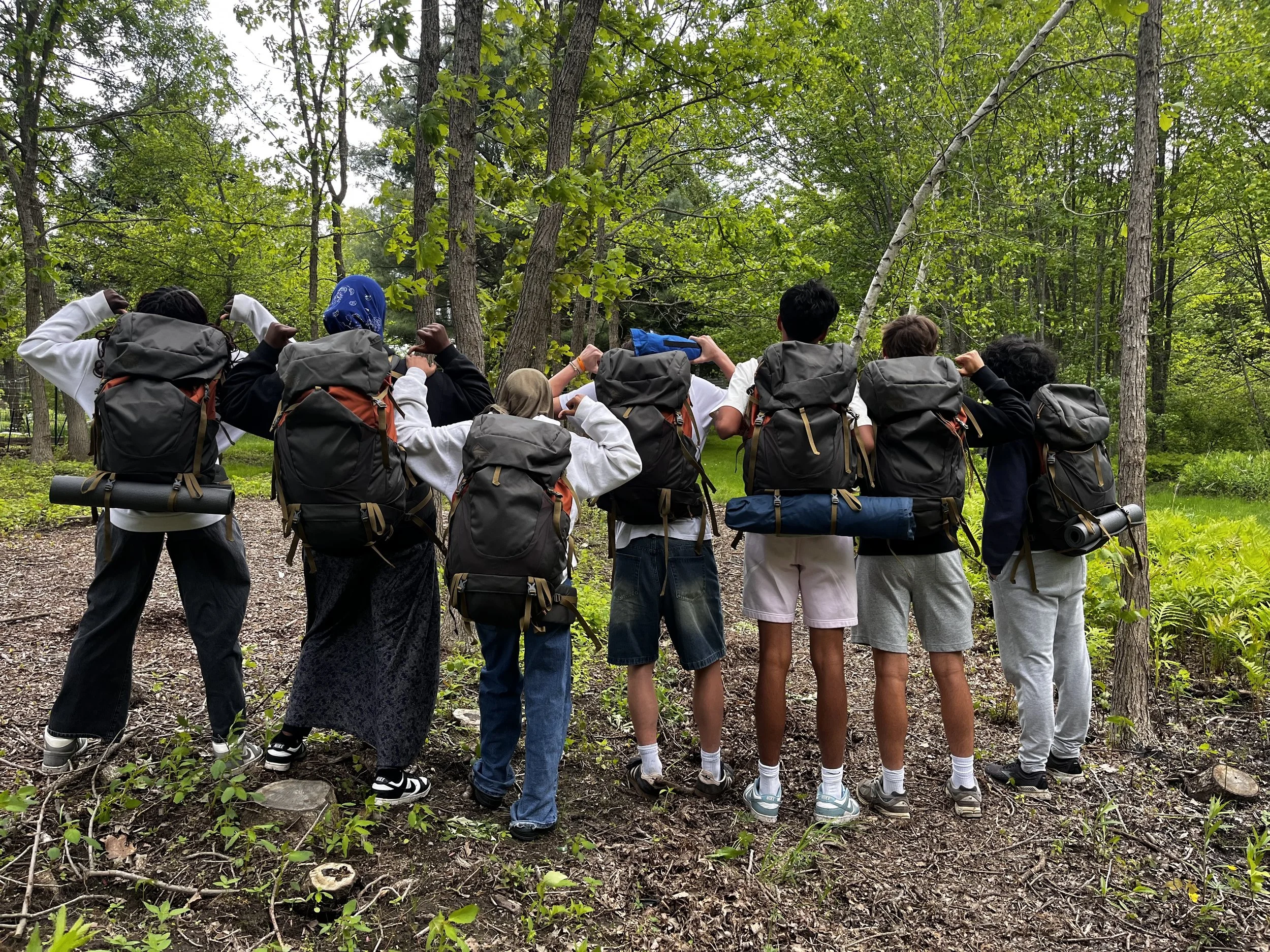 Seven students, two wearing a hijab, pose facing away from the camera with black backpacking backpacks