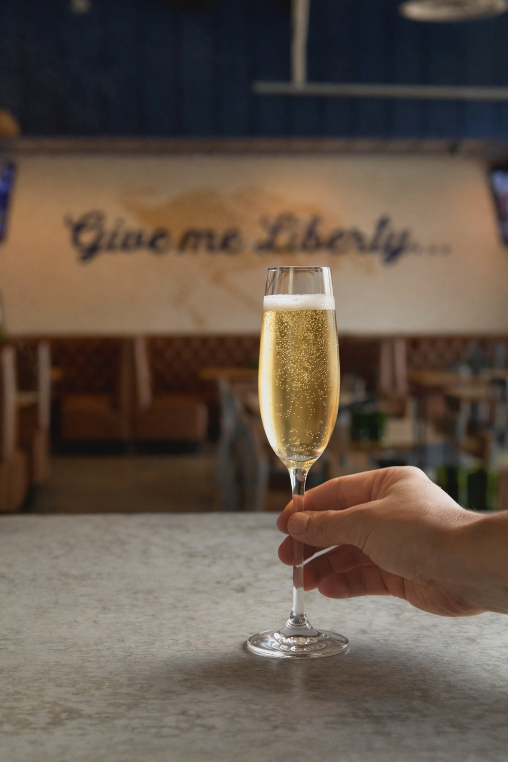 Champagne flute filled with sparkling bubbles held above a marble bar at Liberty Tavern, with the warm dining room and “Give me Liberty” wall signage softly blurred in the background.