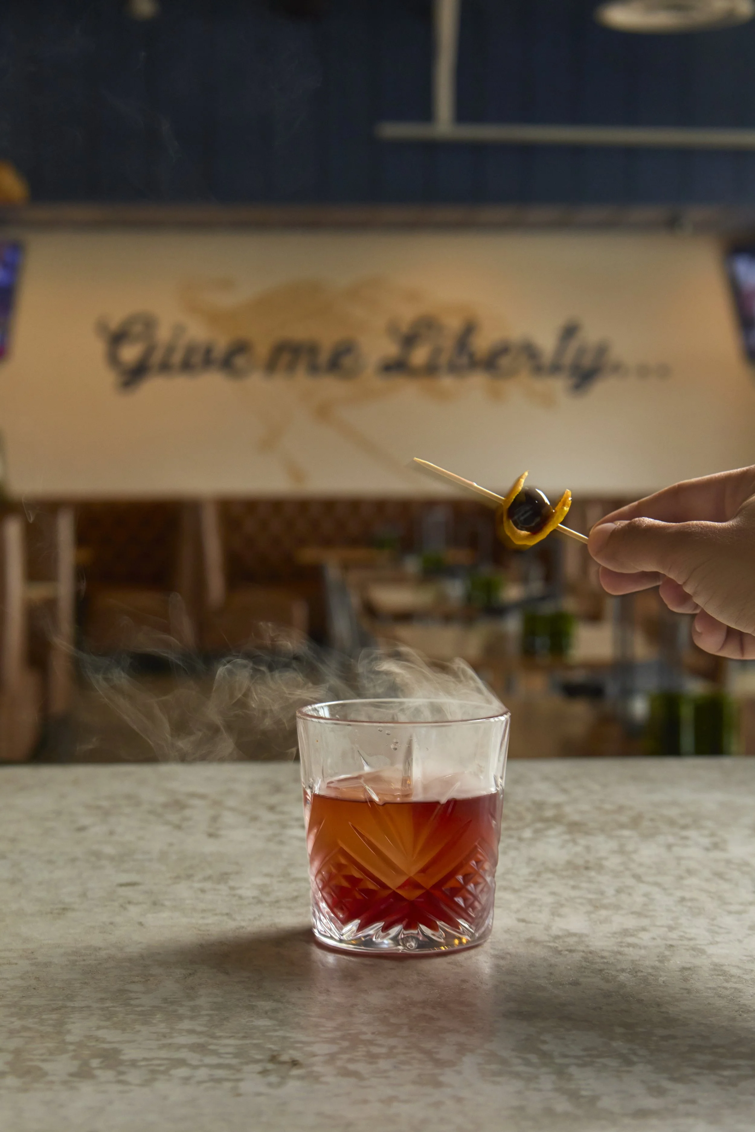 Smoked old fashioned cocktail on a marble bar at Liberty Tavern, garnished with a cherry on a cocktail pick, with smoke rising and the “Give me Liberty…” mural softly blurred in the background.