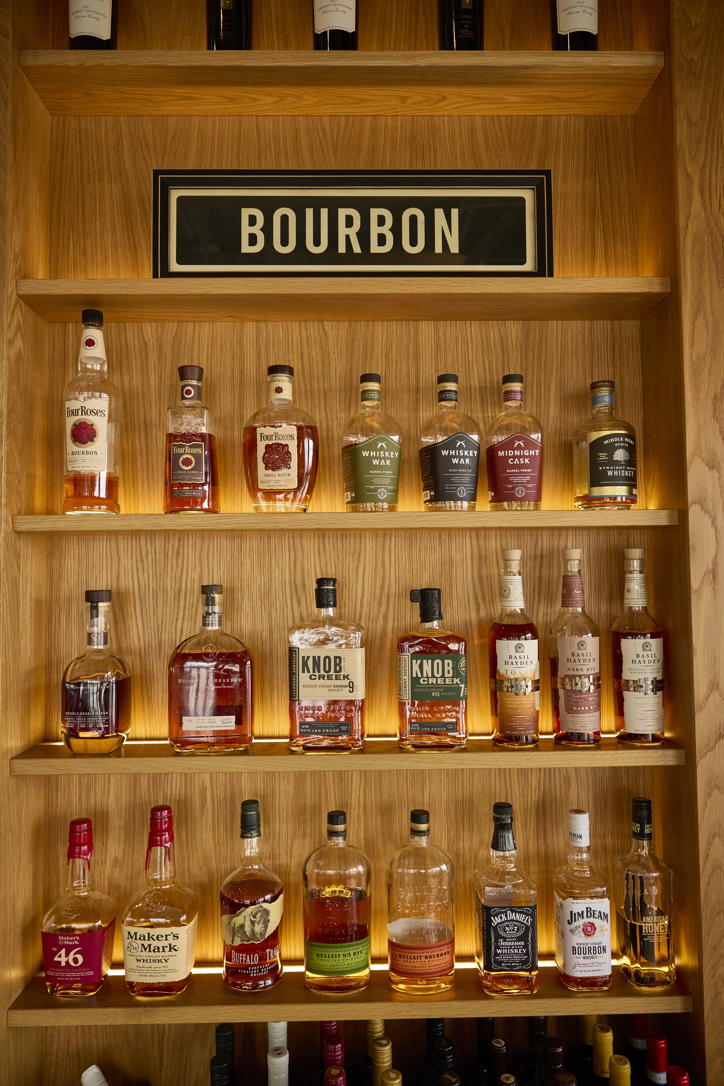 warmly lit wooden shelf displaying a curated selection of bourbon bottles, including Four Roses, Knob Creek, Basil Hayden’s, Maker’s Mark, and more. A bold “BOURBON” sign crowns the top, creating a cozy, refined bar vibe.