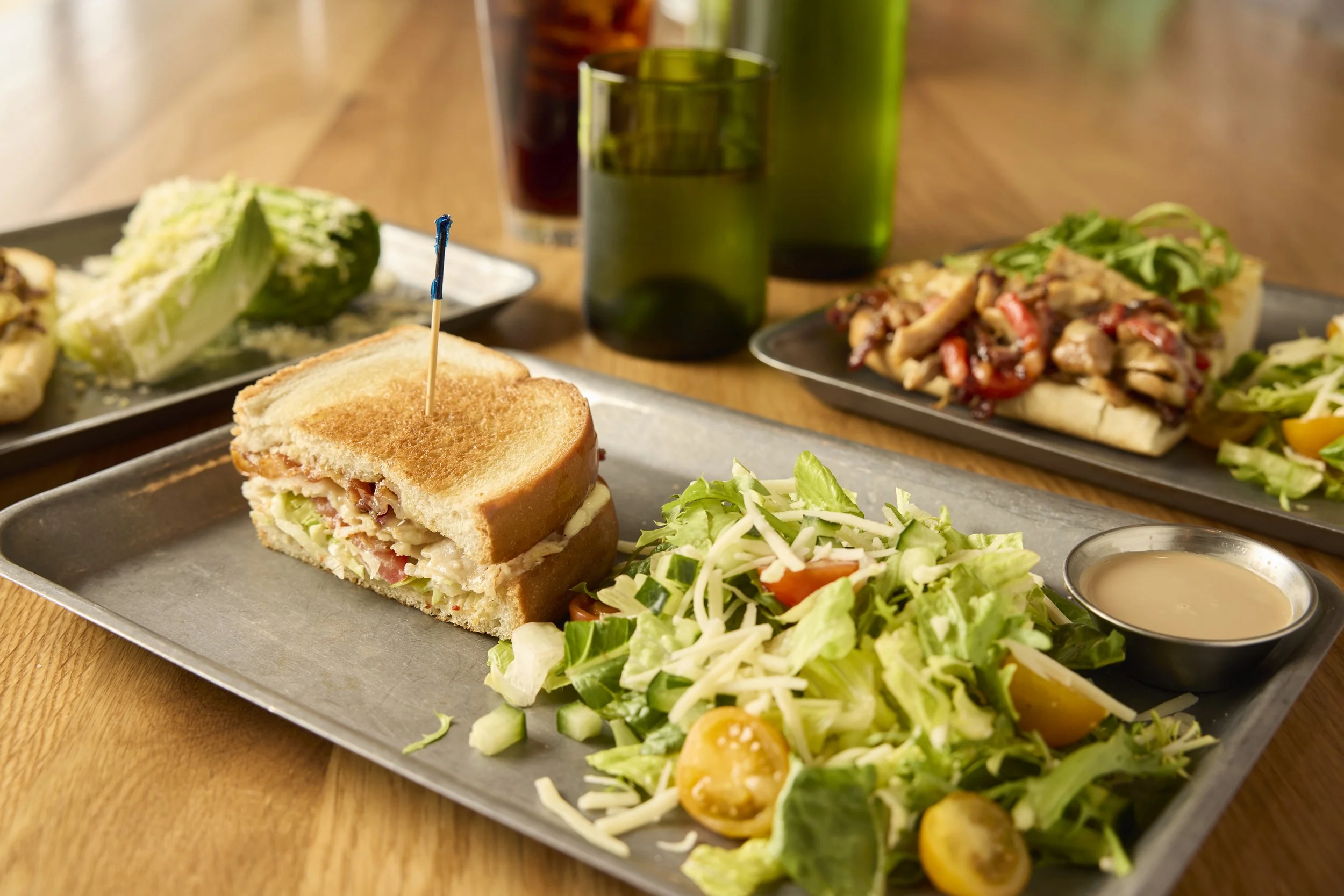 Liberty Tavern lunch spread featuring a toasted club sandwich served with a fresh garden salad and house dressing, with additional sandwiches and drinks on a wood table in the background.