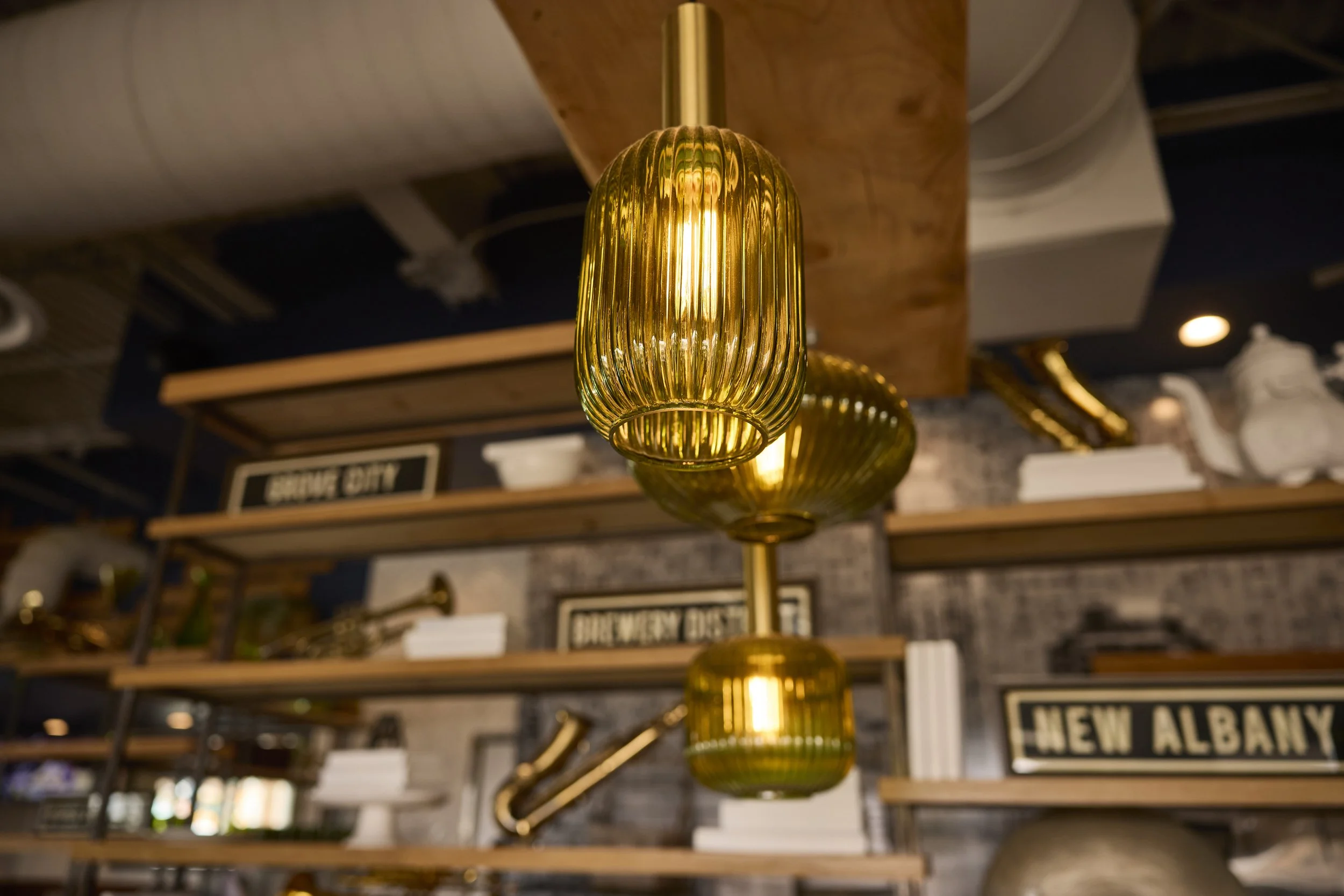 Warm gold pendant lights hanging above wood shelving inside Liberty Tavern, casting a soft glow over vintage decor, brass accents, and framed New Albany signage for an inviting, upscale tavern atmosphere.