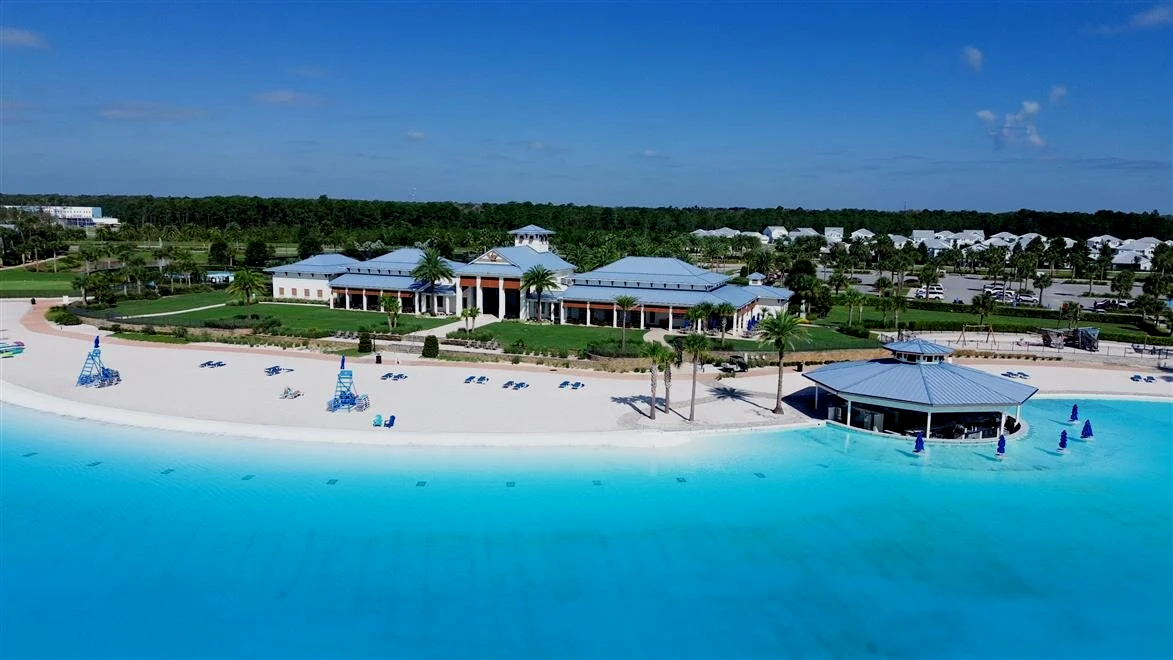 Aerial view of a resort with a beach, pool, and hotel buildings surrounded by palm trees under a blue sky.