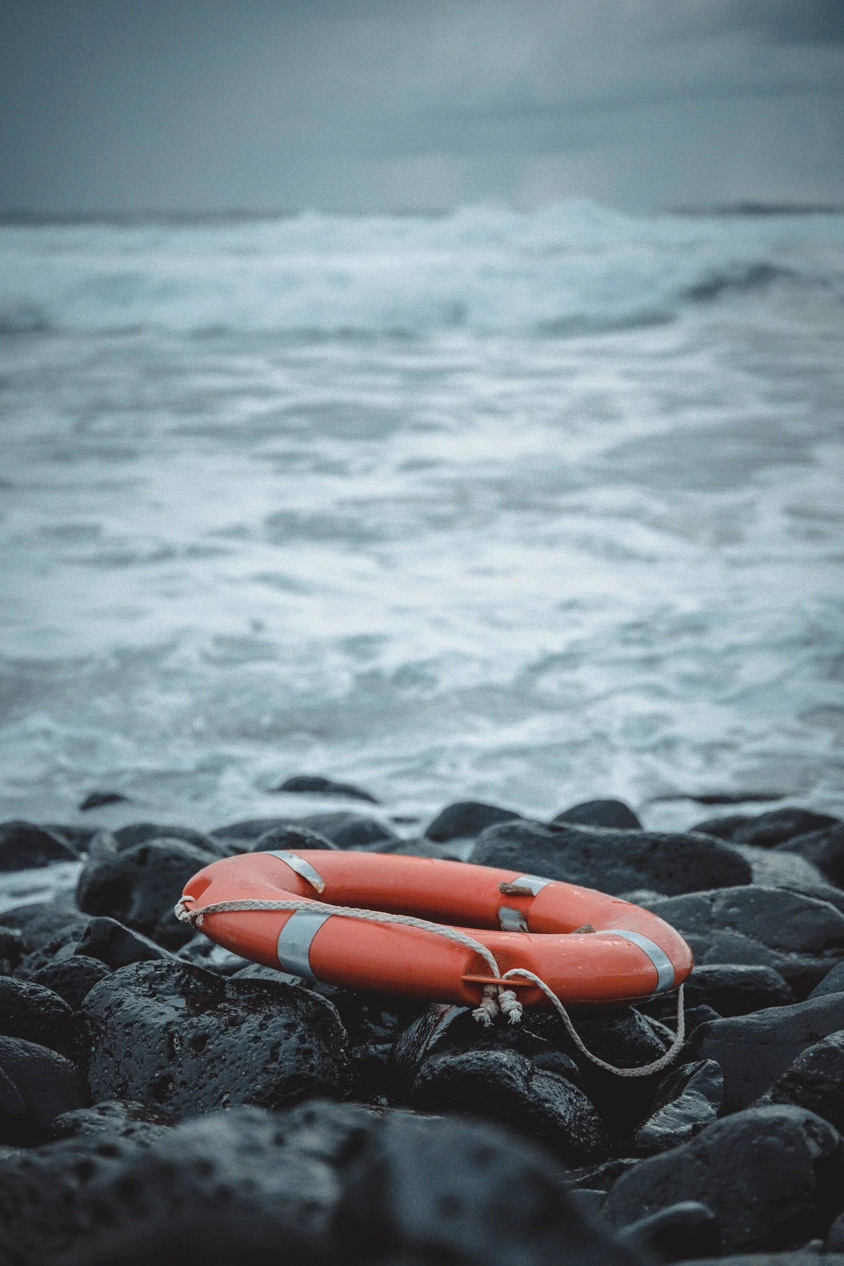 An orange life ring with a rope lies on wet black rocks by the shoreline with waves and a cloudy sky in the background.