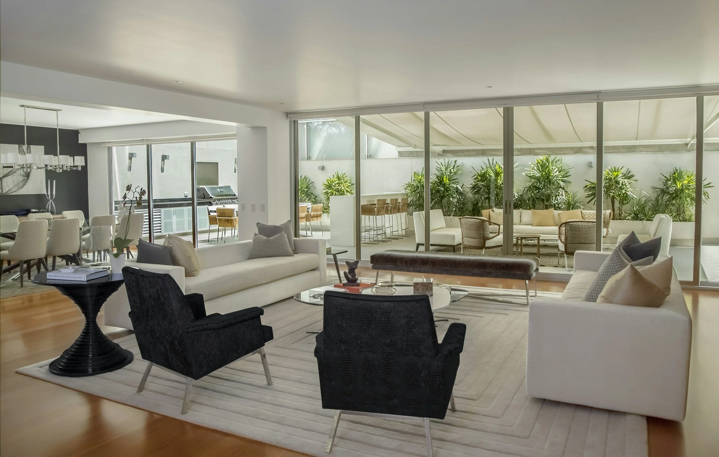 Modern living room with white sofas, black armchairs, glass coffee table, and large glass sliding doors leading to an outdoor patio with lounge chairs and lush green plants.