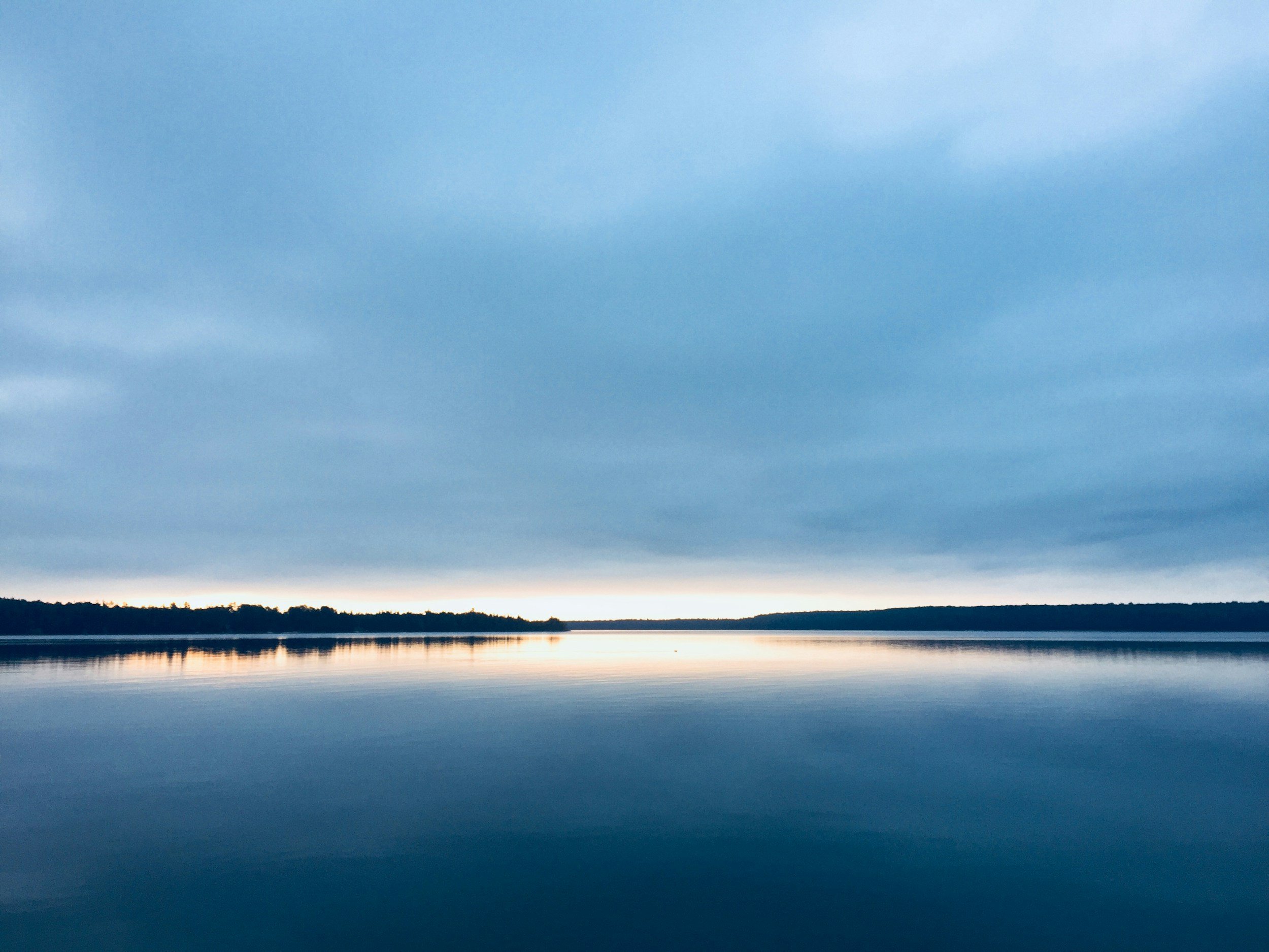 Calm lake with a horizon line of trees against a cloudy sky at sunset or sunrise.