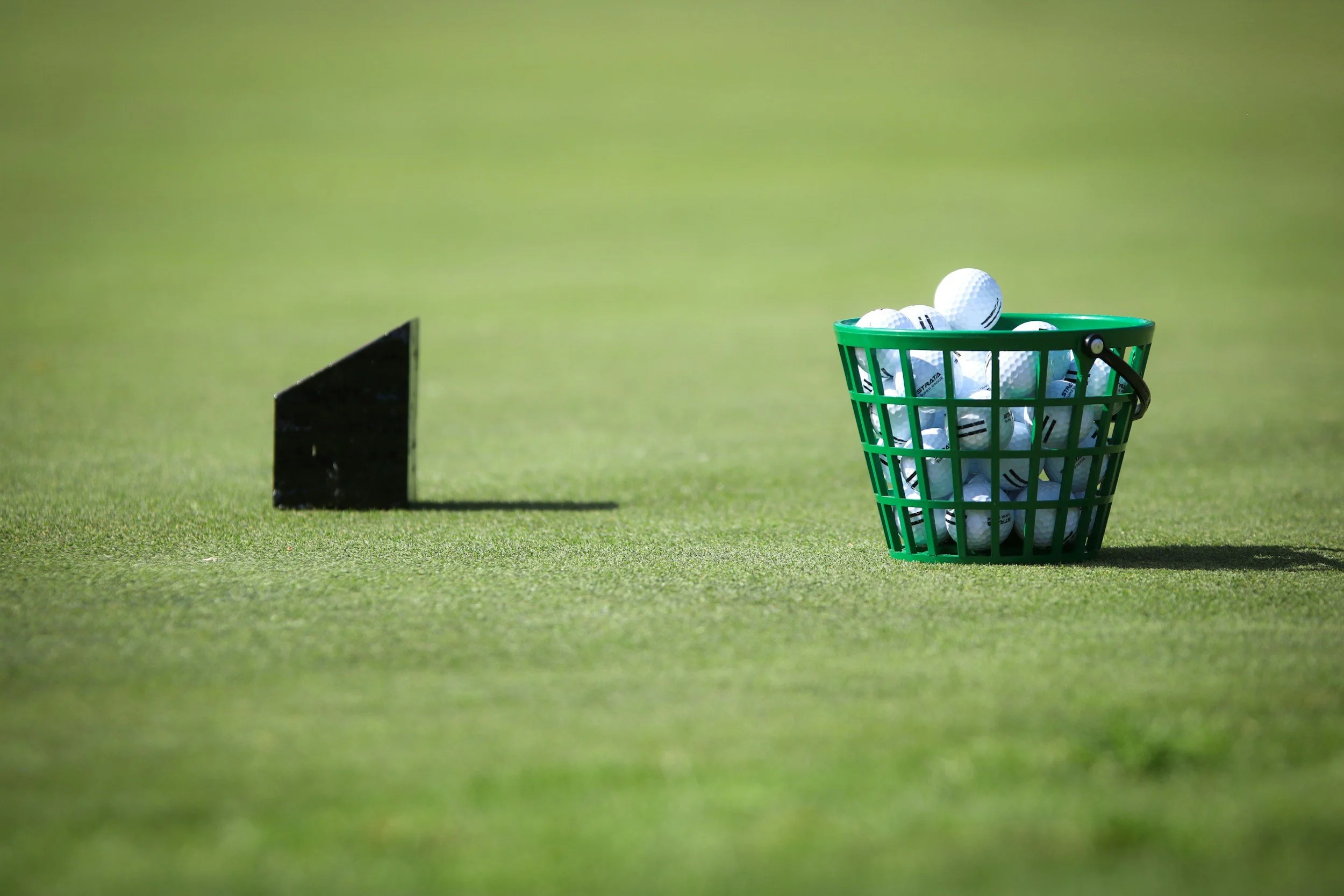 Golf balls in a green basket on a golf course, with a black golf tee marker nearby, on a well-manicured grass field.