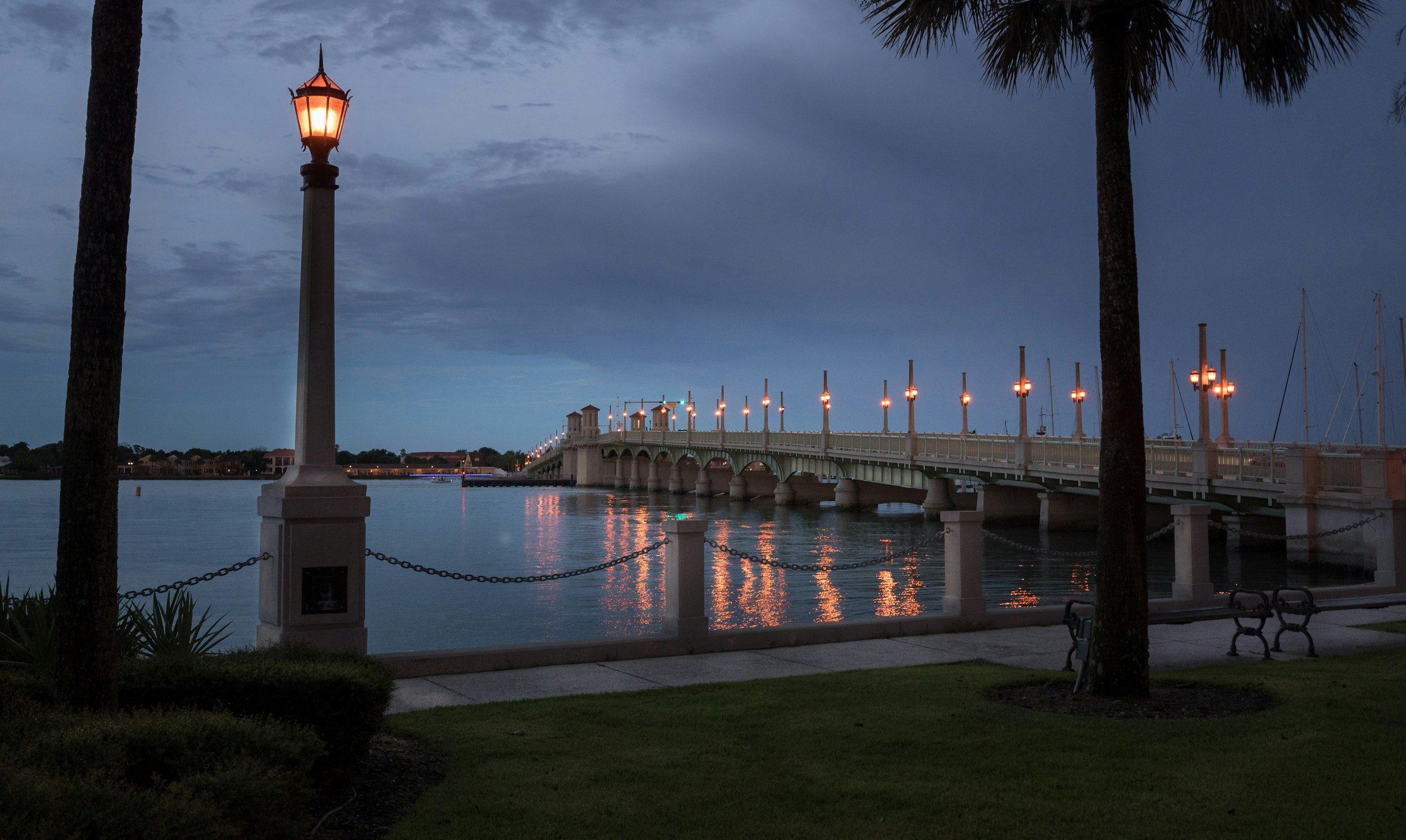 A waterfront scene at dusk featuring a bridge with illuminated lampposts, calm water reflecting the lights, and trees and benches in the foreground.