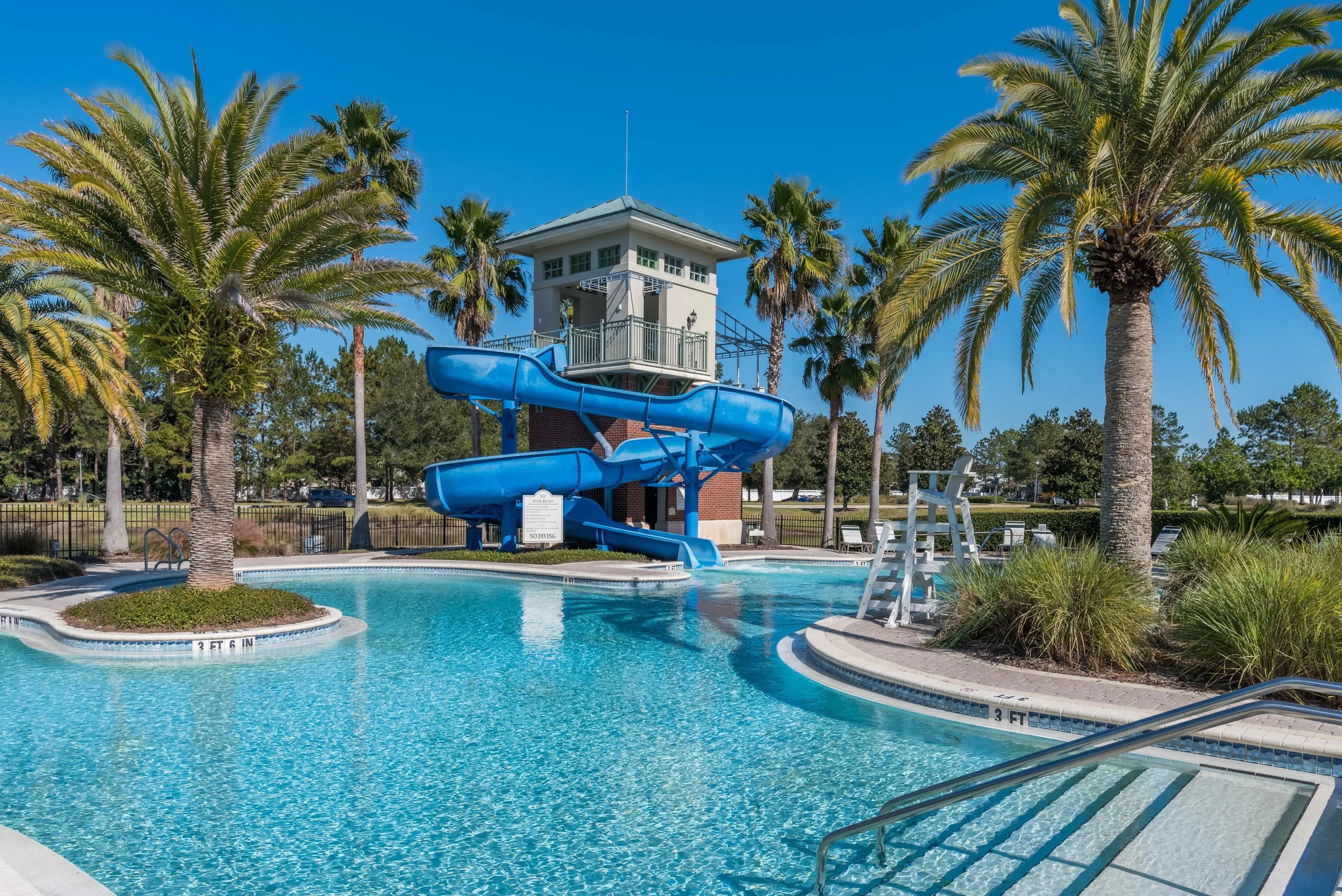 A swimming pool with a blue water slide surrounded by palm trees and a tall pool tower under a clear blue sky.