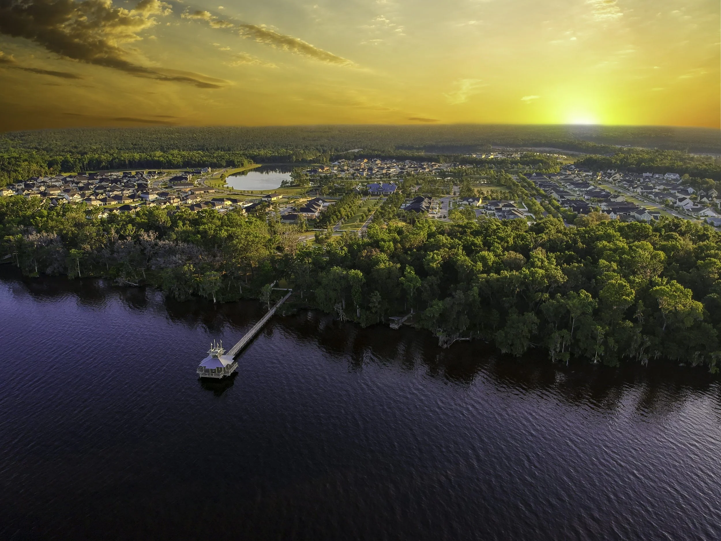 An aerial view of a neighborhood near a body of water during sunset, with trees, houses, and a dock extending into the water.