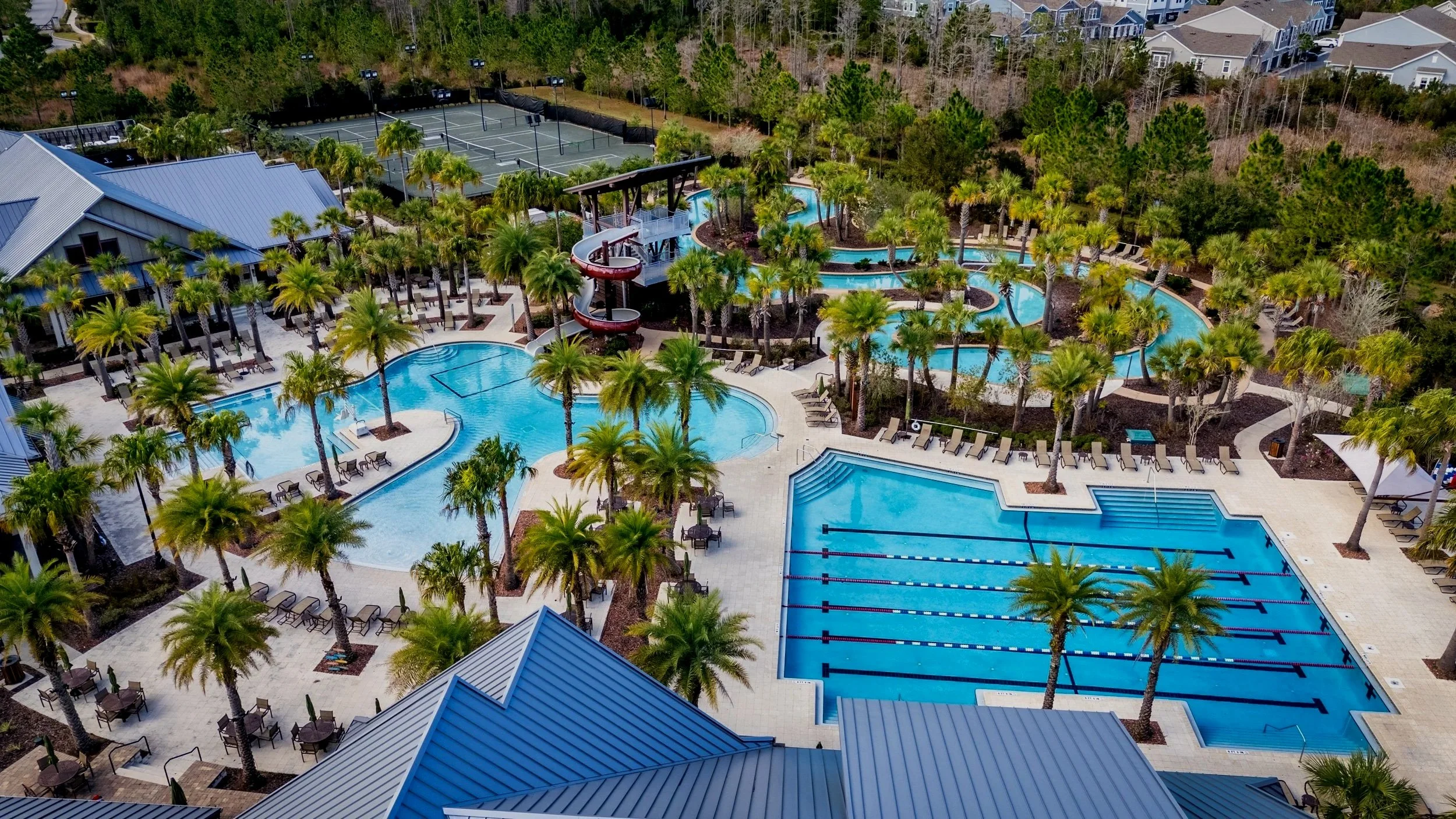 Aerial view of a resort pool area with multiple pools, a waterslide, surrounded by numerous palm trees, lounge chairs, and a nearby building with a metal roof. Tennis courts are visible in the background, along with a wooded area.