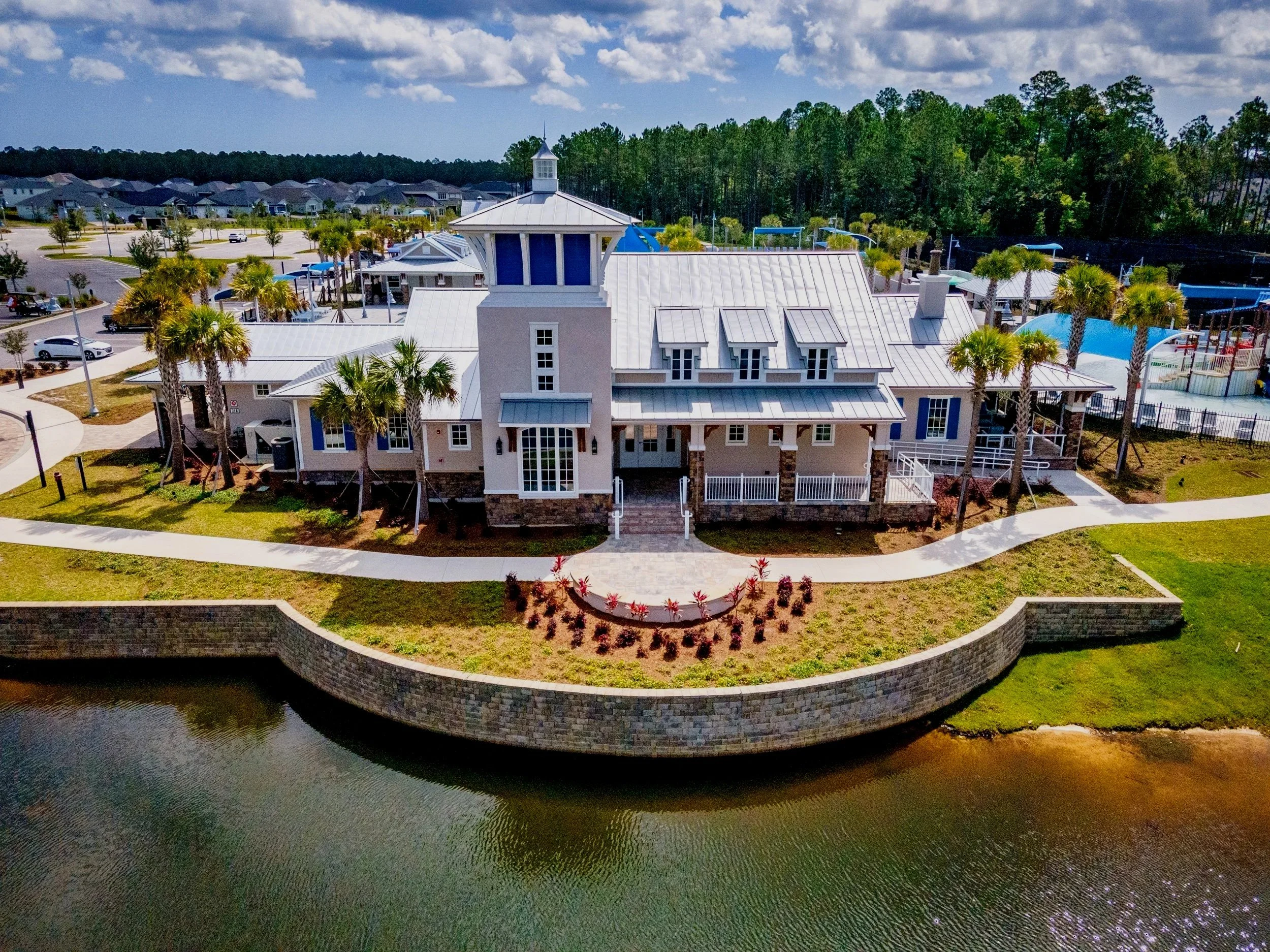 Aerial view of a large house with multiple gabled roofs, white exterior, and a tower, situated by a water body, surrounded by landscaped greenery, palm trees, and a curved walkway.