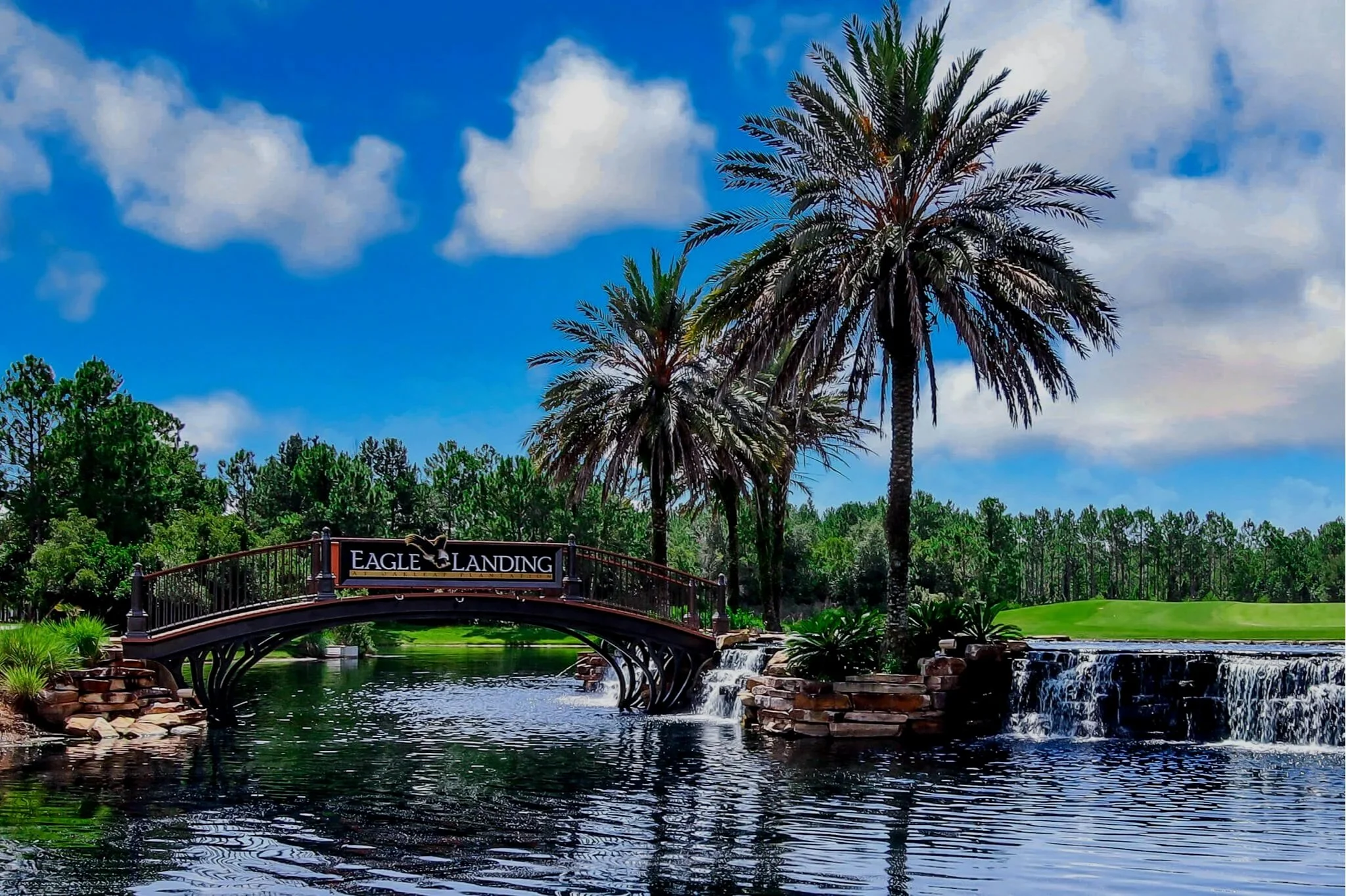 A scenic view of a bridge labeled 'Eagle Landing' over a pond with waterfalls, surrounded by palm trees and lush greenery under a partly cloudy sky.