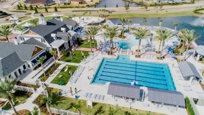 A community pool area with a large swimming pool, surrounded by palm trees, lounge chairs, and poolside structures, with residential houses and a waterway in the background.