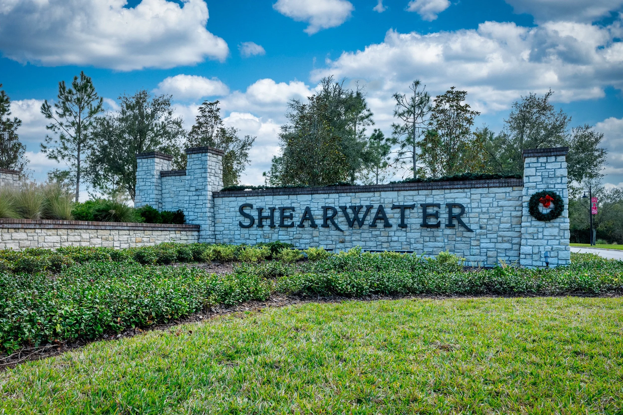 Photo of a large stone sign that reads 'SHEARWATER' with a wreath decoration, set against a backdrop of trees and a partly cloudy sky.
