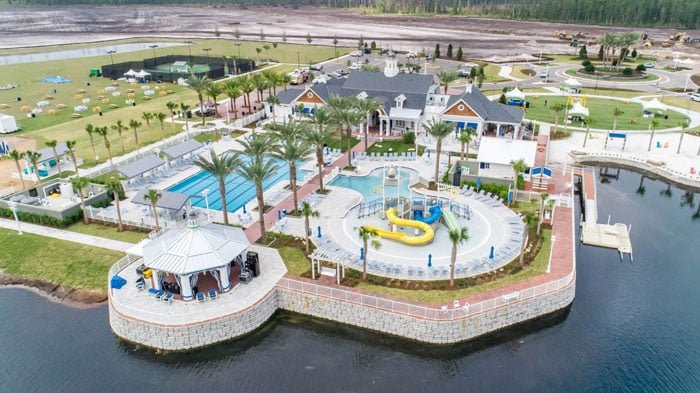 Aerial view of a lakeside recreational area with pools, a waterslide, a gazebo, palm trees, and surrounding lawns.