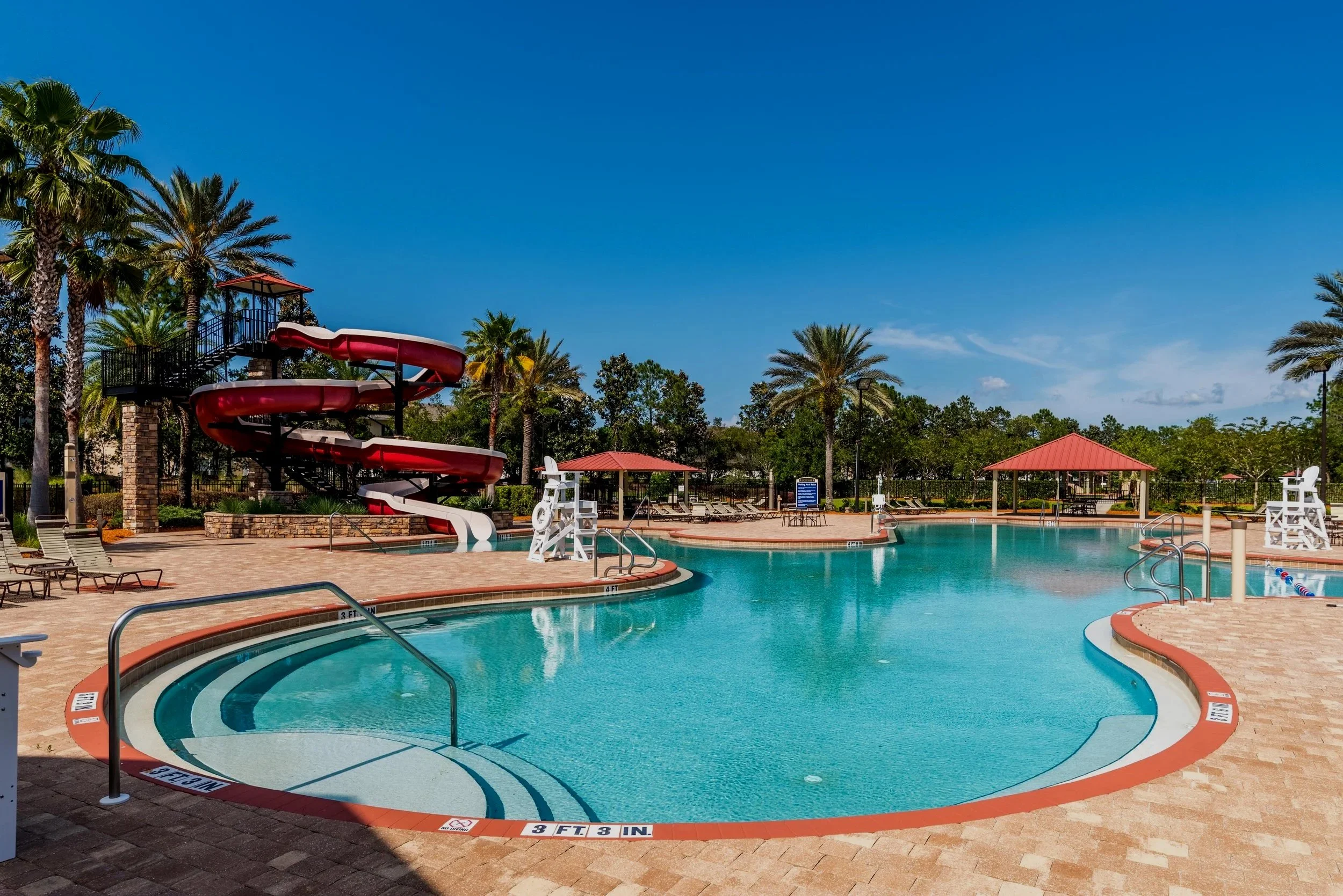 Empty outdoor swimming pool with a red water slide, surrounded by palm trees and lounge chairs, under a blue sky.
