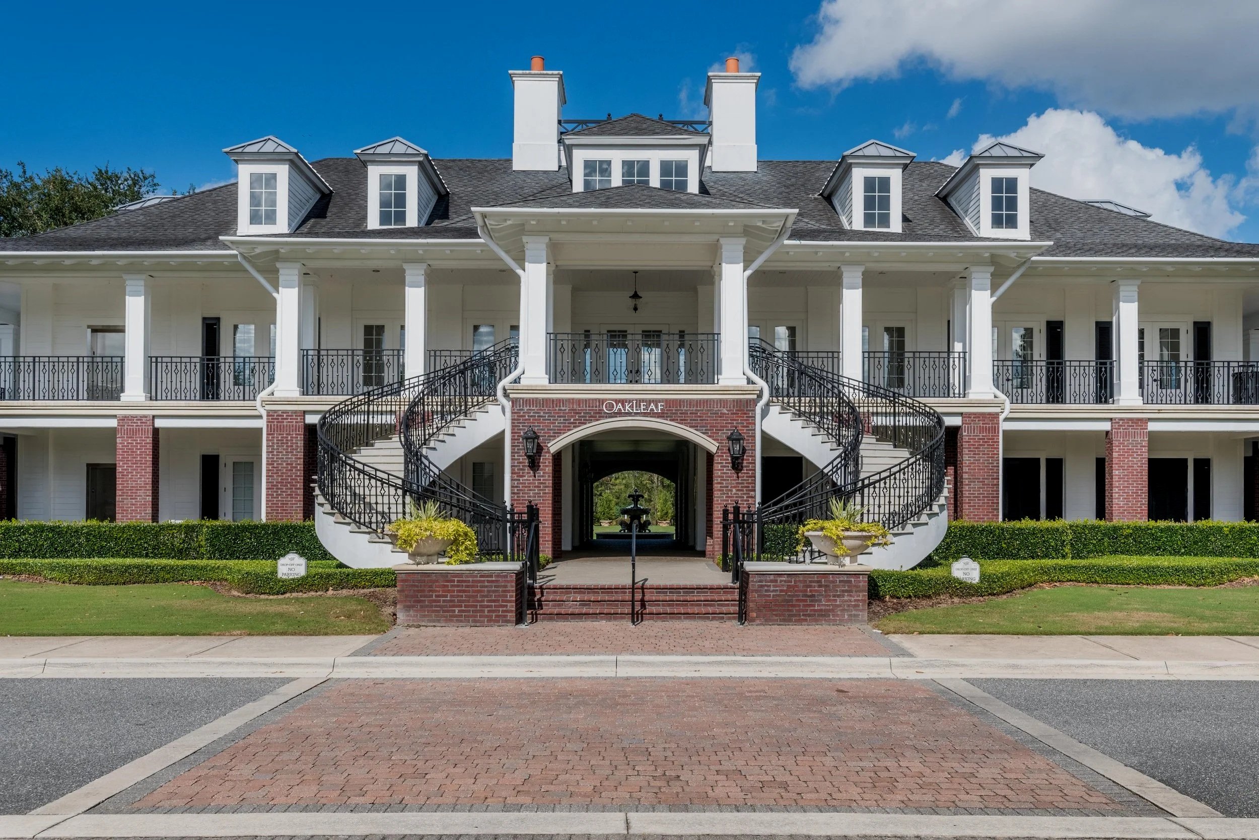 Front view of a large, white, multi-story residential building with two curved staircases leading to the main entrance, black railings, red brick accents, and a small fountain visible through the archway under the entrance. The building is named Oakleaf.