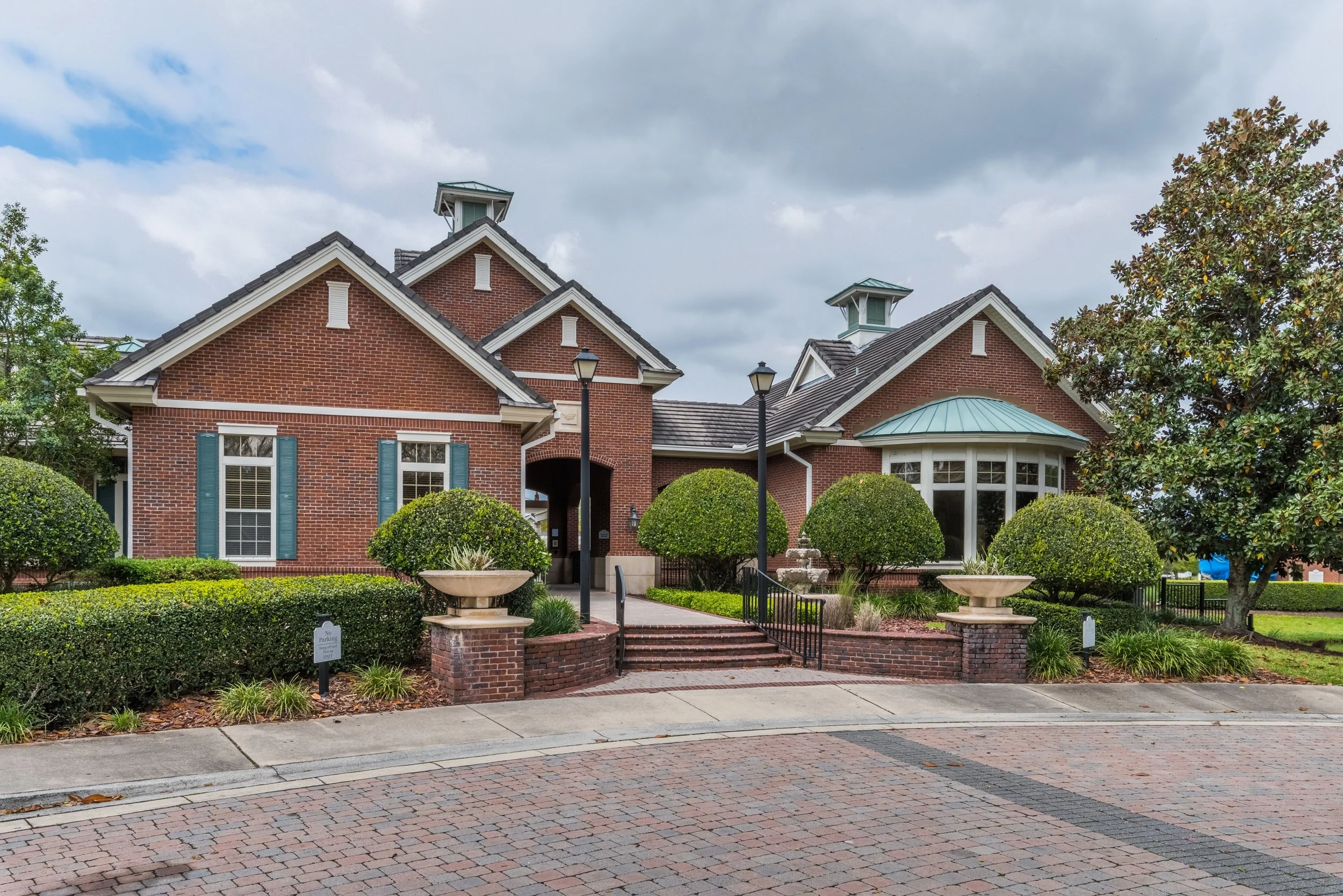 A large brick house with a landscaped front yard featuring trimmed bushes, planters, and trees under a cloudy sky.