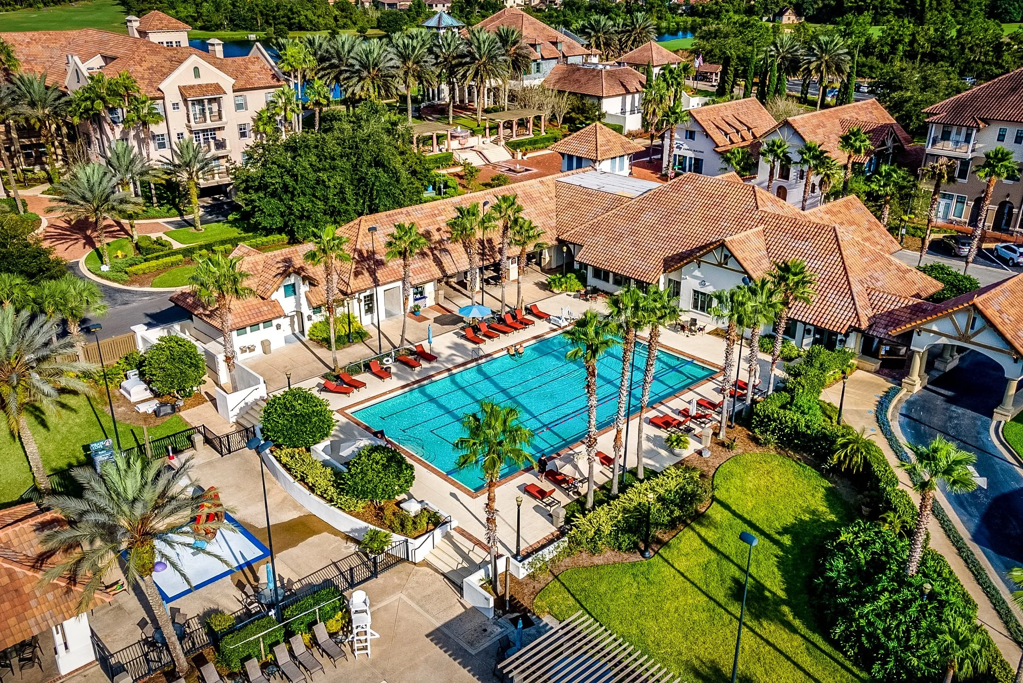 Aerial view of a resort with a large swimming pool, surrounded by palm trees, lounge chairs, and surrounding buildings with red-tile roofs.