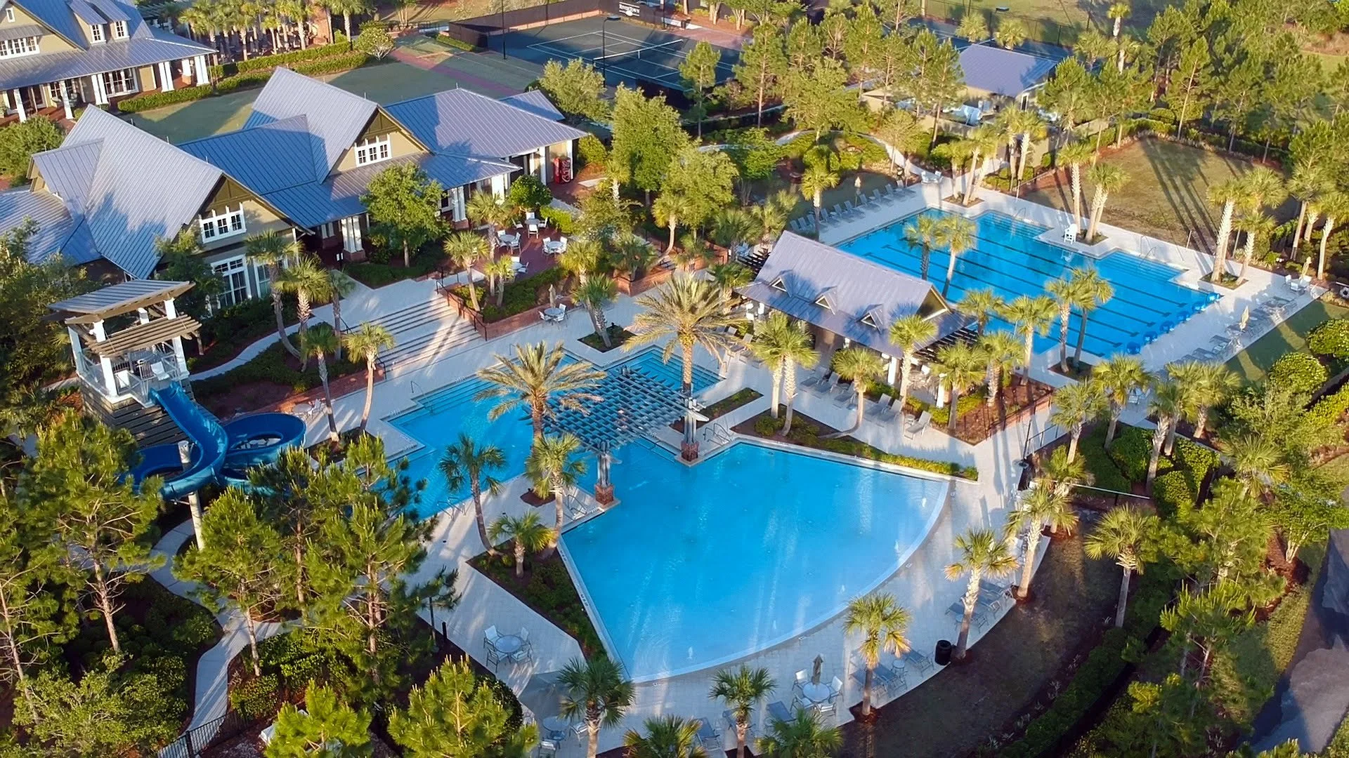 Aerial view of a resort-style complex with multiple swimming pools, a waterslide, tennis courts, and surrounding buildings, lush green trees, and paved walkways.