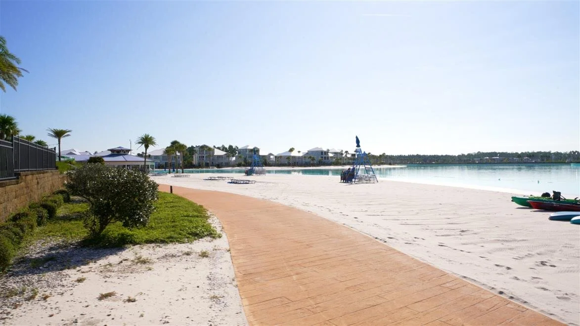 Sunny beach with a pathway, paddleboats, lifeguard chairs, and beachfront houses in the distance.