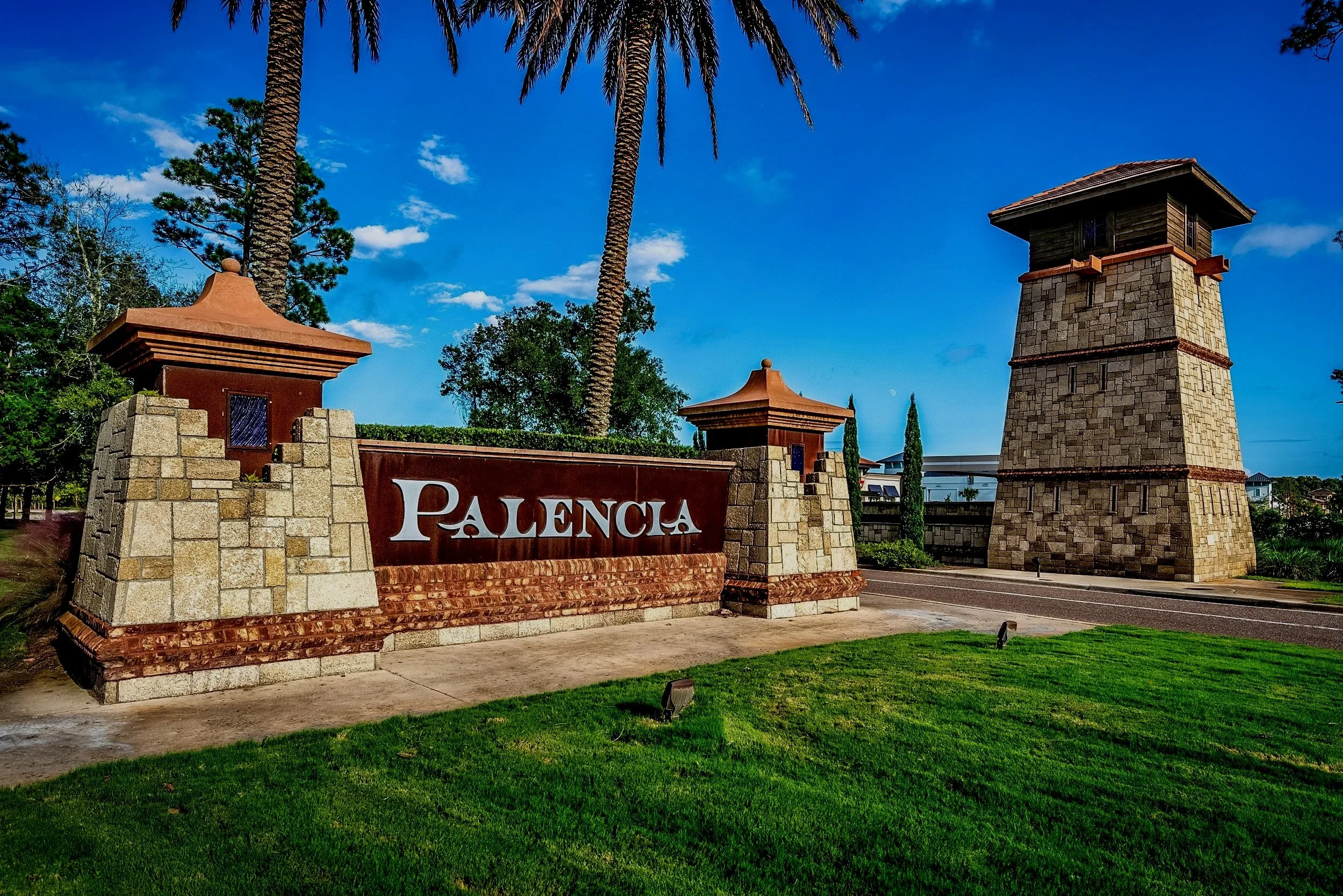 Entrance sign for Palencia with stone pillars and a large stone tower in the background, surrounded by green grass and tall palm trees under a blue sky with some clouds.