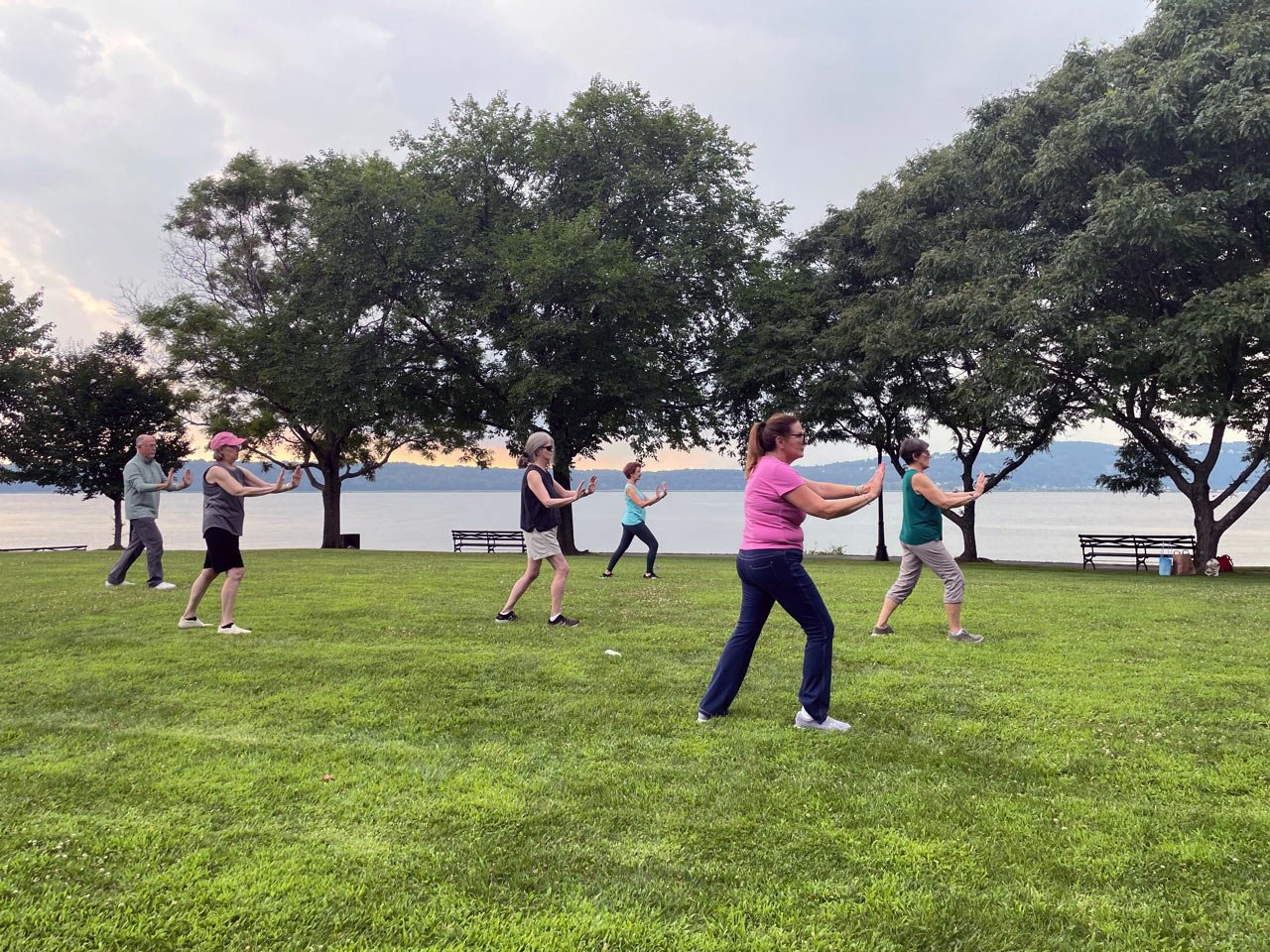 A group of six adults practicing Tai Chi in a park near a lake, with trees and benches in the background.