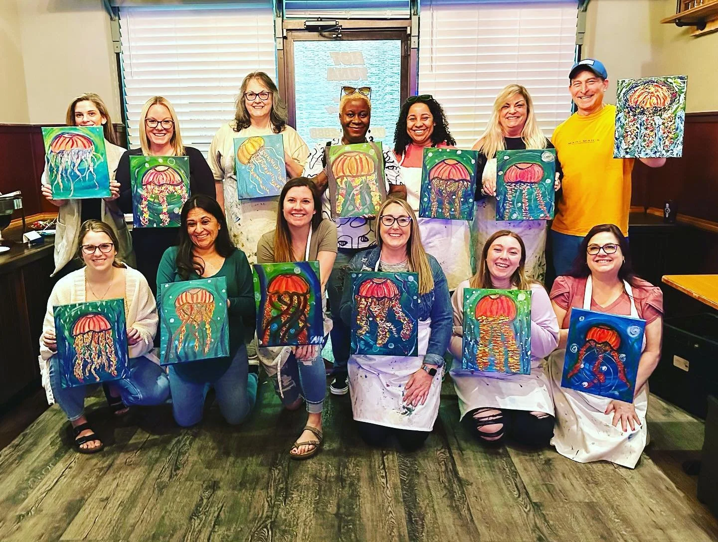 A group of people at a paint and sip event in Virginia Beach holding up paintings of jellyfish they created, smiling in a room with wooden floors and blinds.