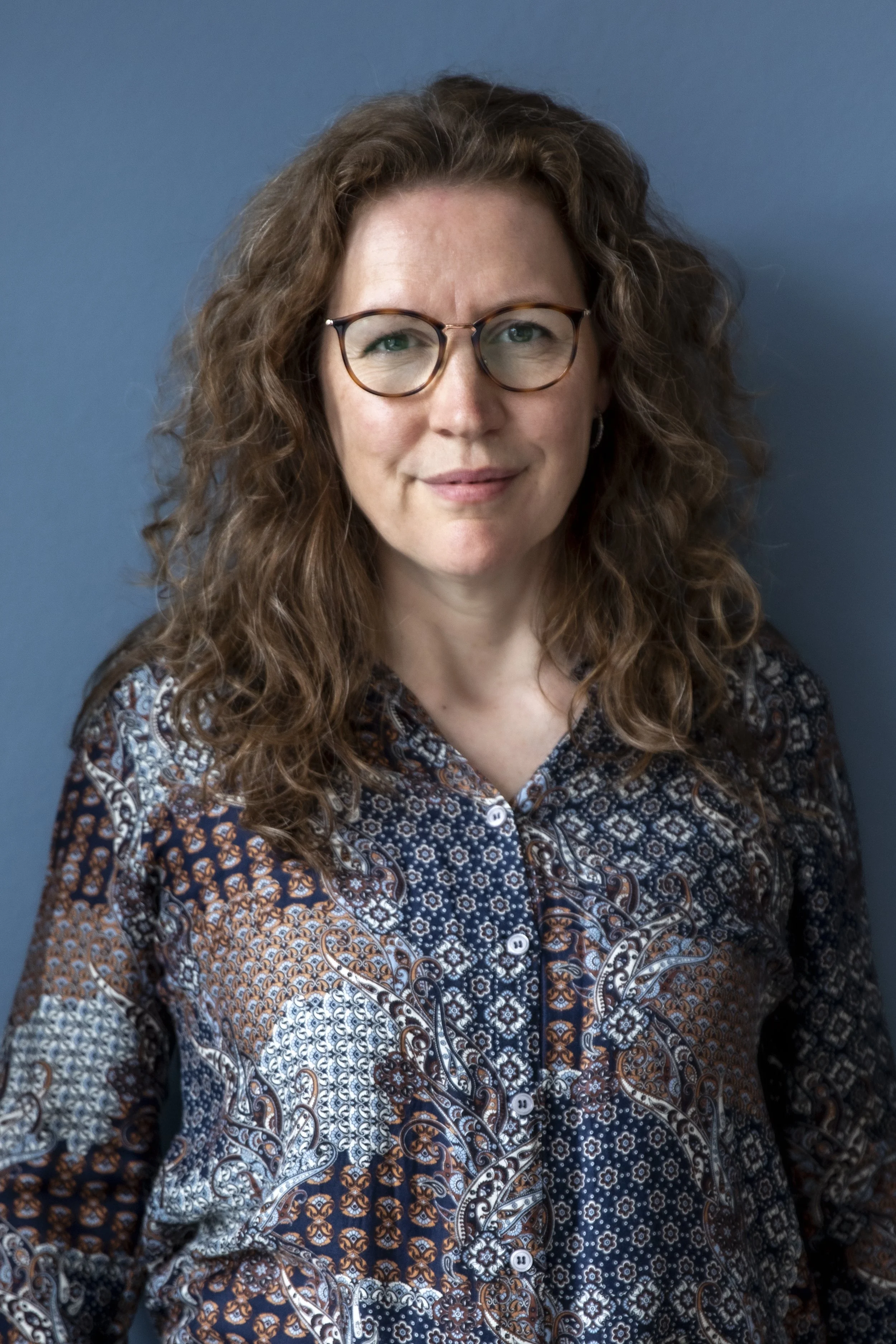 Portræt af en kvinde med brunt, krøllet hår og briller, iført en mønstret bluse, stående mod en blå væg.