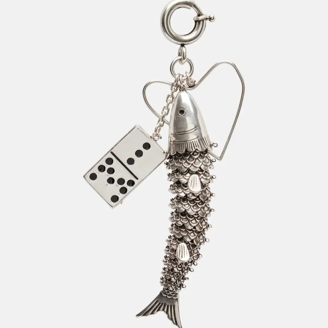 Silver fish-shaped keychain with a domed head, textured scales, and a fin on the side, attached to a heart-shaped wire, with a domino piece and a small chain hanging from a metal ring.