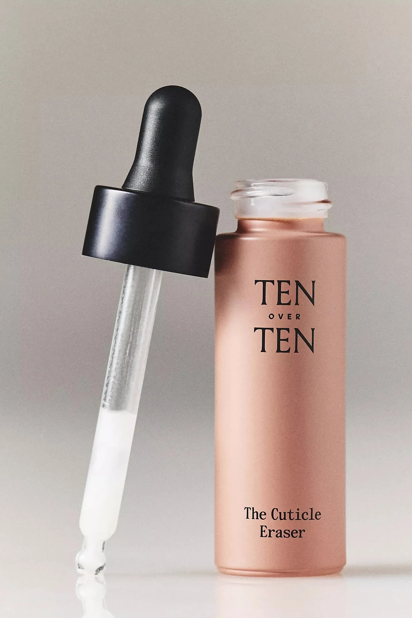 A skincare product with a dropper and a pink container labeled 'TEN OVER TEN The Cuticle Eraser' on a neutral background.