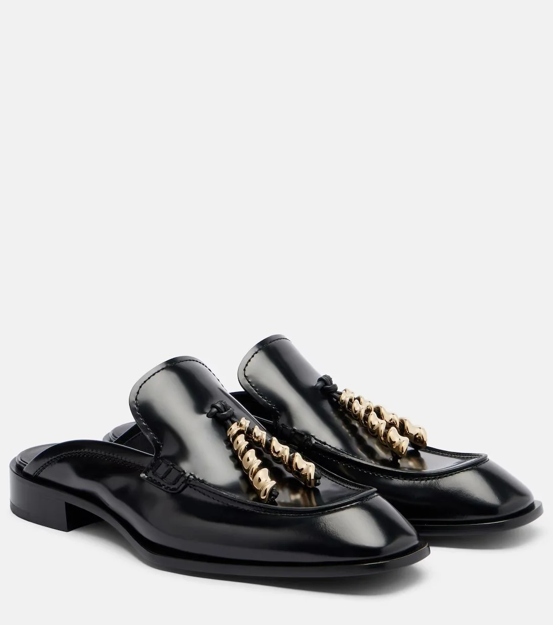 Pair of shiny black leather loafers with decorative gold chain and black tassel detail on the laces, isolated on white background.