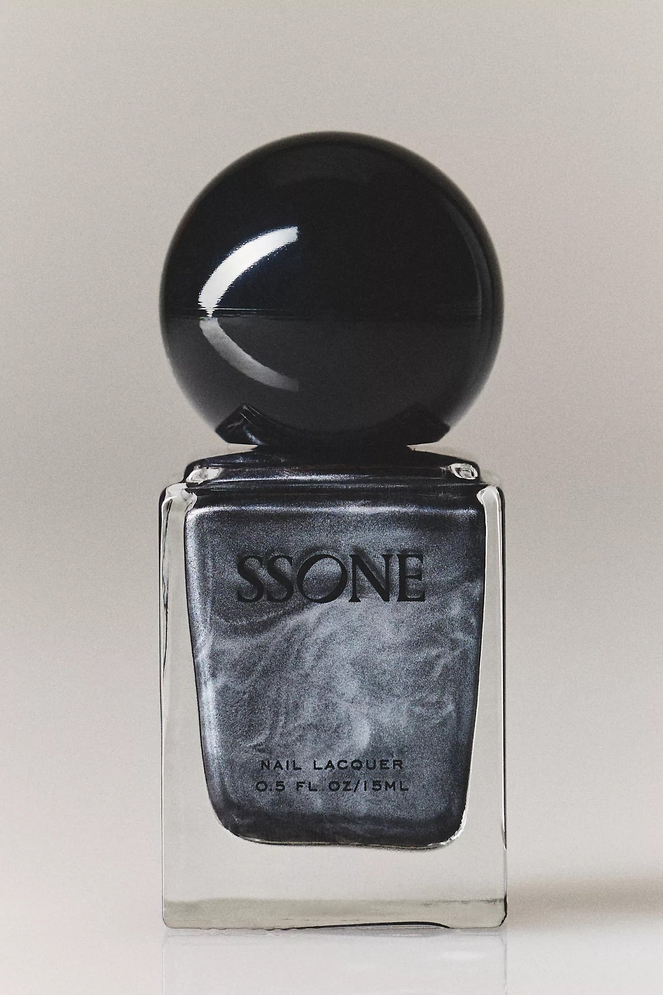 A bottle of SSONE nail lacquer in a black and silver shiny container with a black spherical cap, placed on a reflective surface against a plain background.