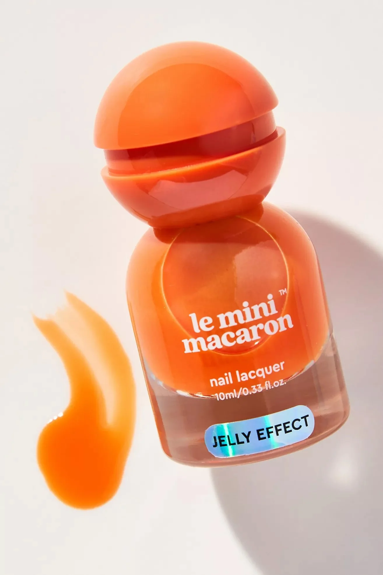 Orange nail lacquers shaped like miniature macaroons with the label "le mini macaron" and the words "Jelly Effect" on the bottle, placed on a white surface with a small swirl of orange nail polish beside them.