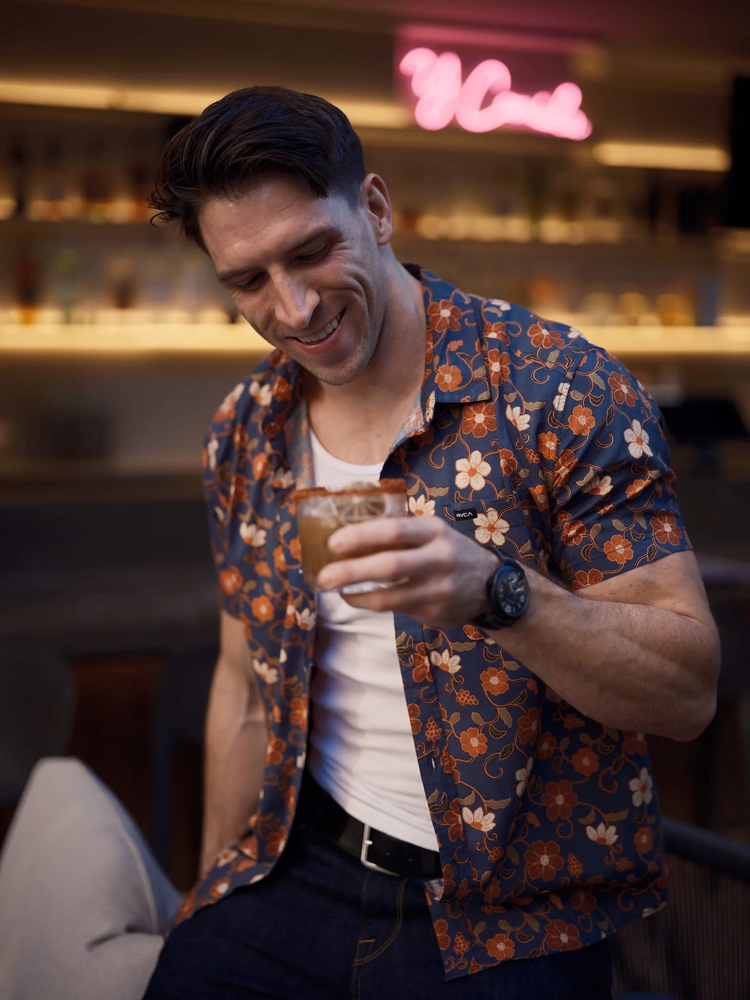 A smiling man in a floral shirt holding a cocktail in a bar setting with neon pink sign in the background.