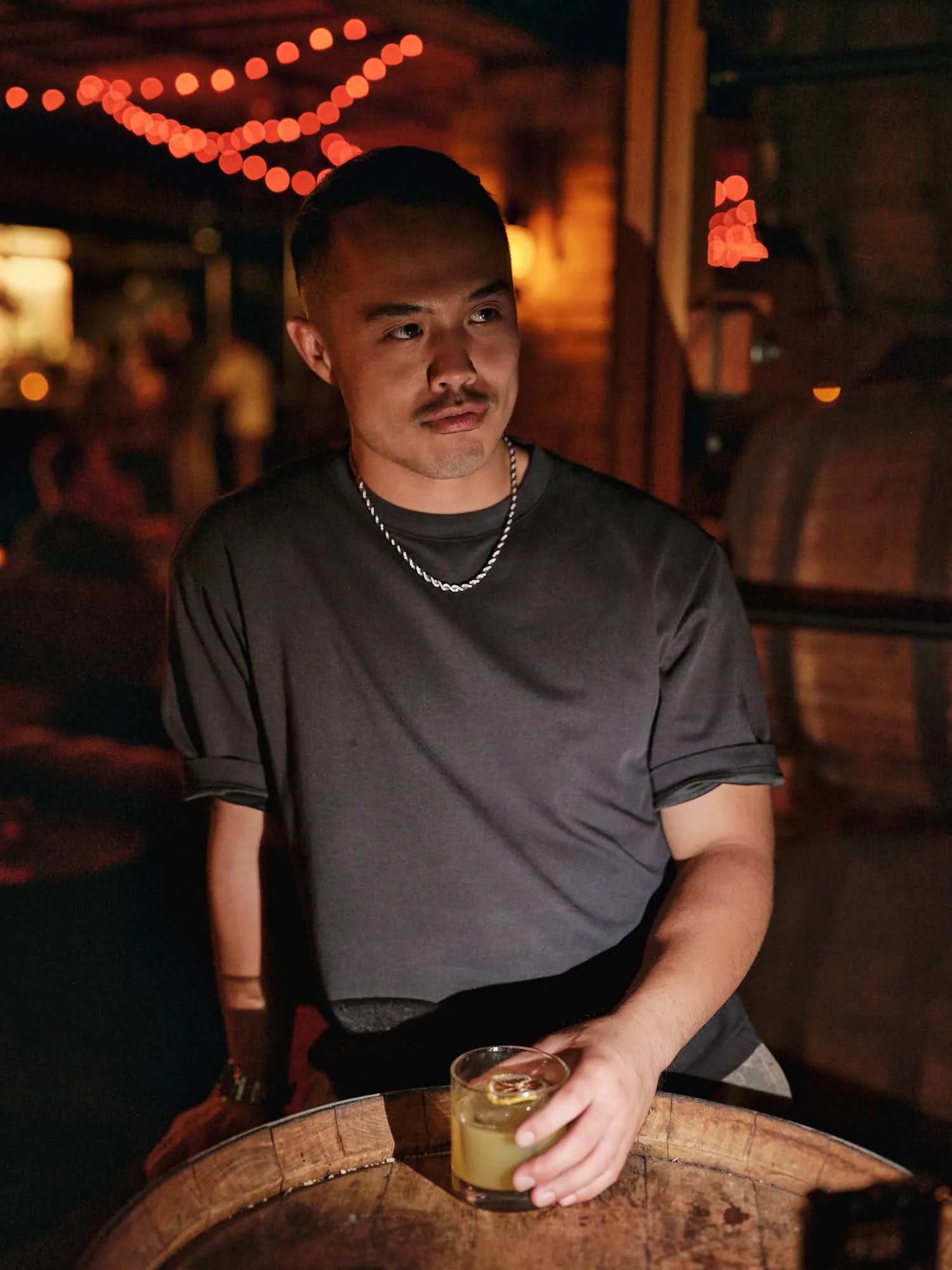 Portrait of a man in a dimly lit bar holding a cocktail beside a wooden barrel, with warm string lights glowing in the background.