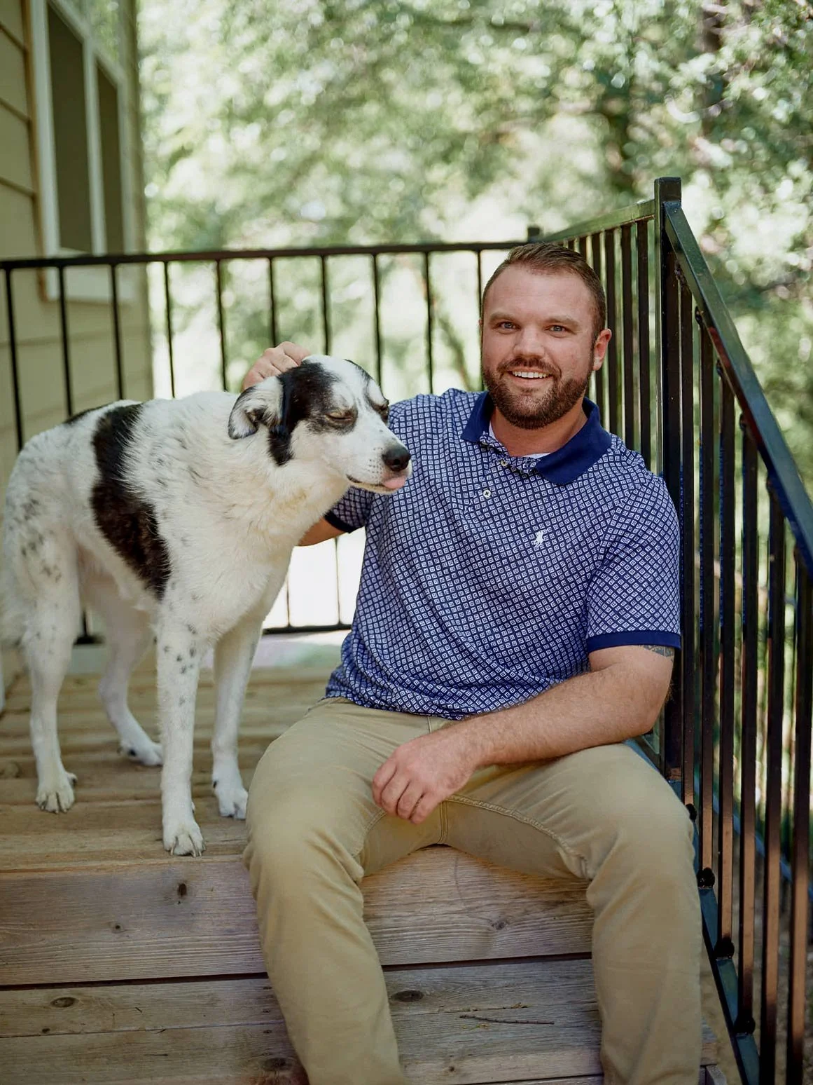 Portrait of a man sitting on wooden steps beside a black-and-white dog, with leafy trees and soft natural light in the background.