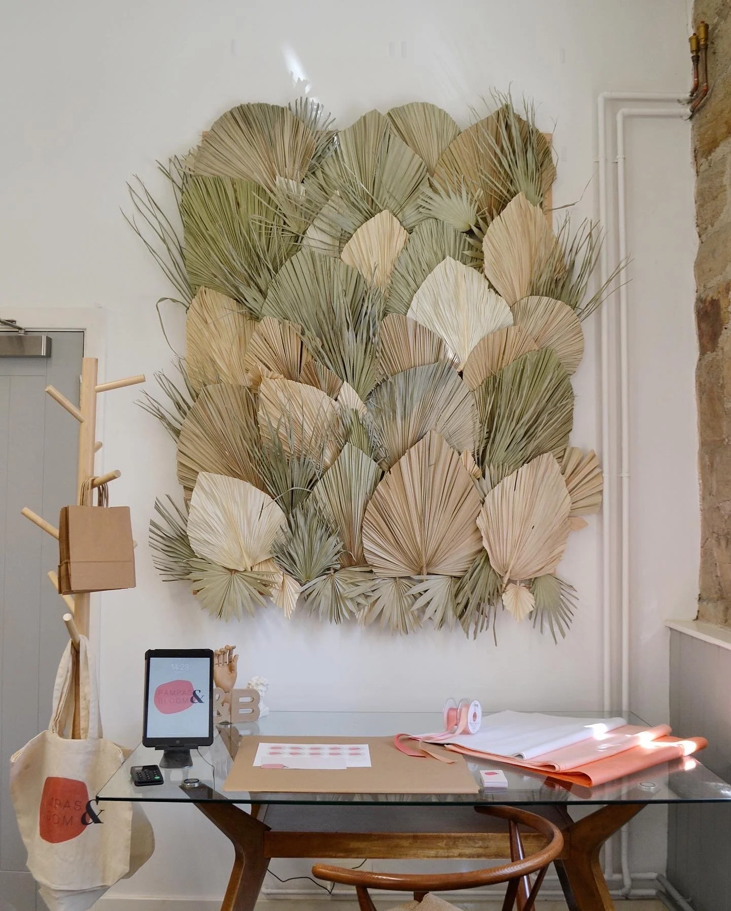 Interior of a retail space with a large wall decoration of dried palm leaves in neutral tones, a wooden table with pink and white paper items, a tote bag, a tablet, and a small coffee table.