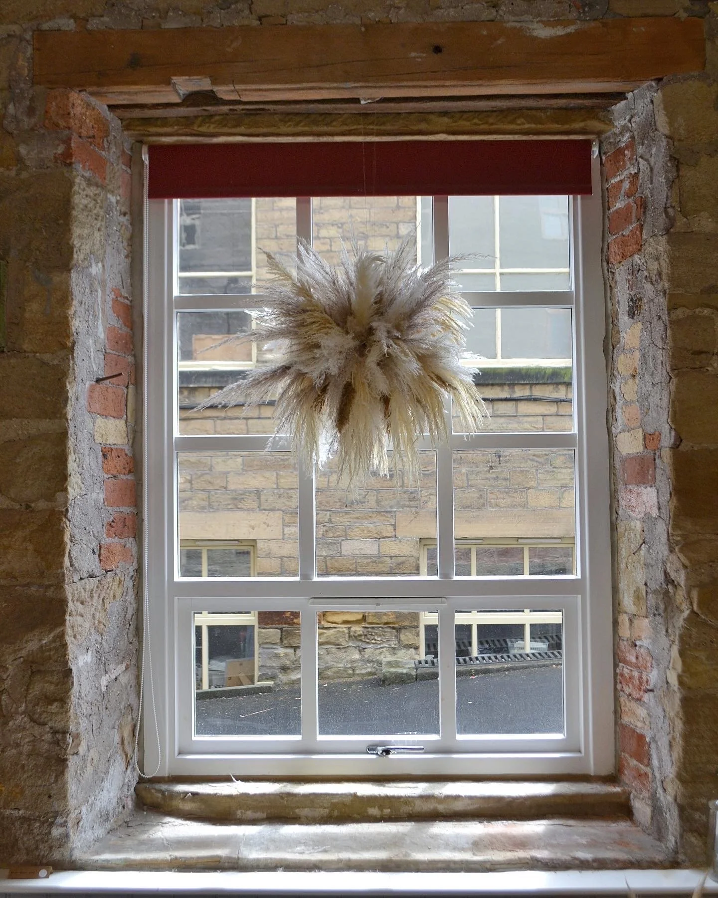 A window with a decorative hanging plant made of pampas grass in the middle, set in a brick wall with stone framing, and a red roller blind partially rolled up at the top.