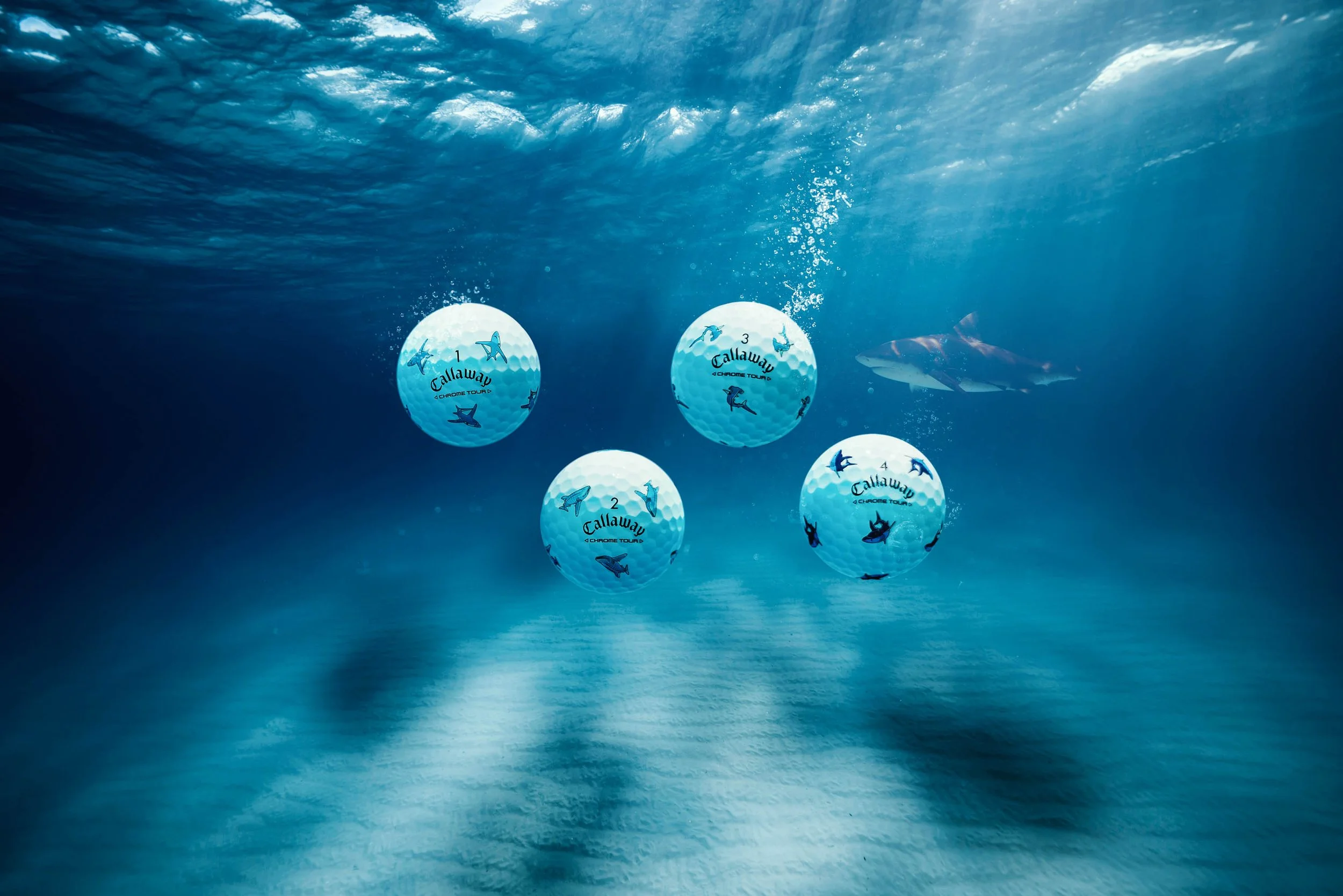 Underwater view of four golf balls with Callaway branding, numbered 1 through 4, floating in a blue ocean with a shark swimming nearby and sunlight filtering through the water surface.