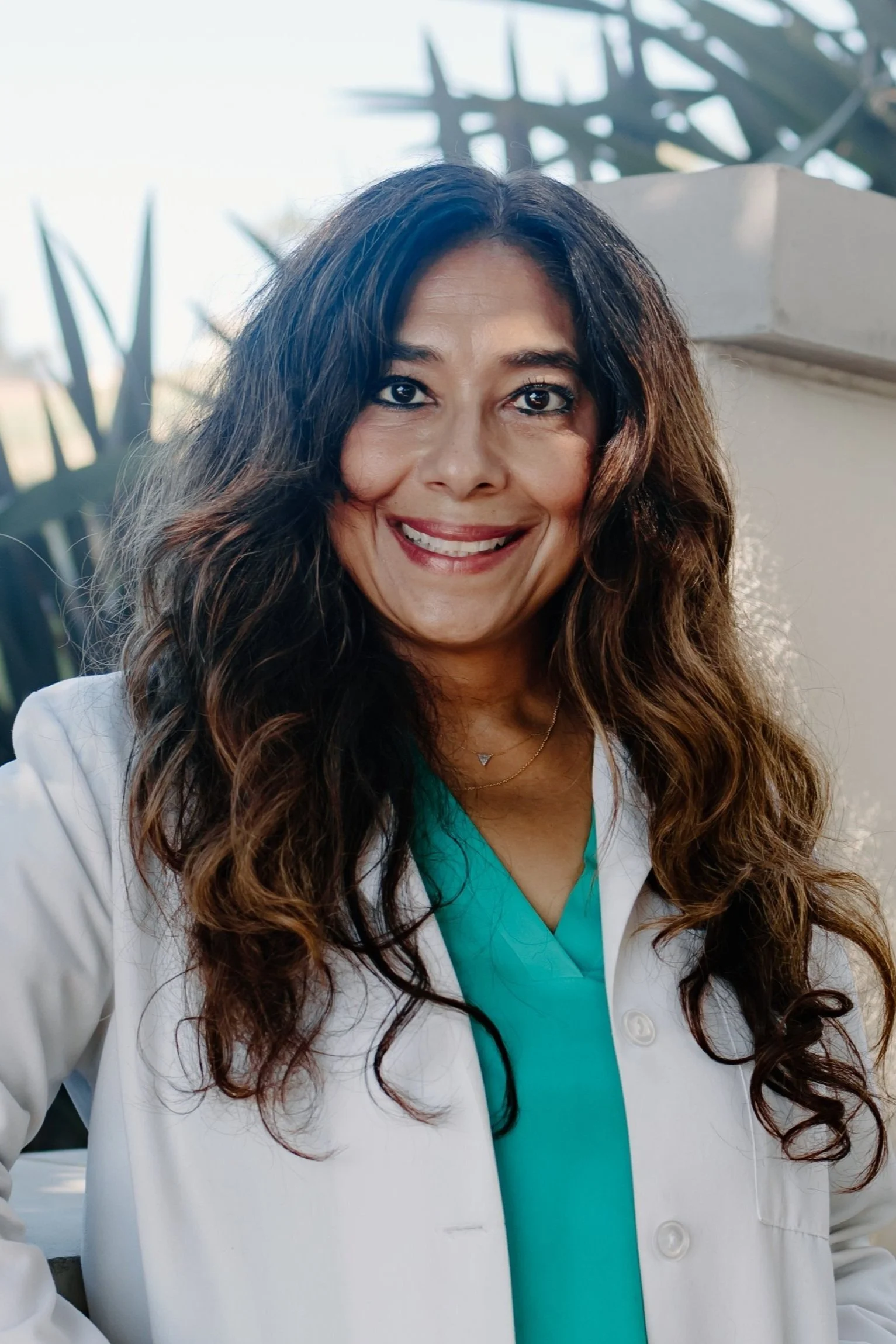 A smiling female healthcare professional with long wavy hair, wearing a white lab coat over teal scrubs, standing outdoors with plants and a light-colored structure in the background.