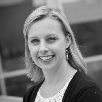 A black and white portrait of a woman with shoulder-length hair, smiling, wearing earrings, a light-colored blouse, and a dark cardigan.