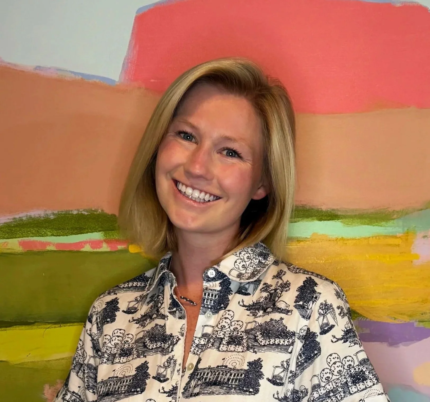 A smiling woman with blonde hair standing in front of a colorful, abstract painted background.