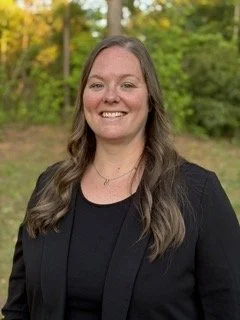 A woman with long brown hair smiling outdoors, wearing a black blazer and top, with green trees in the background.