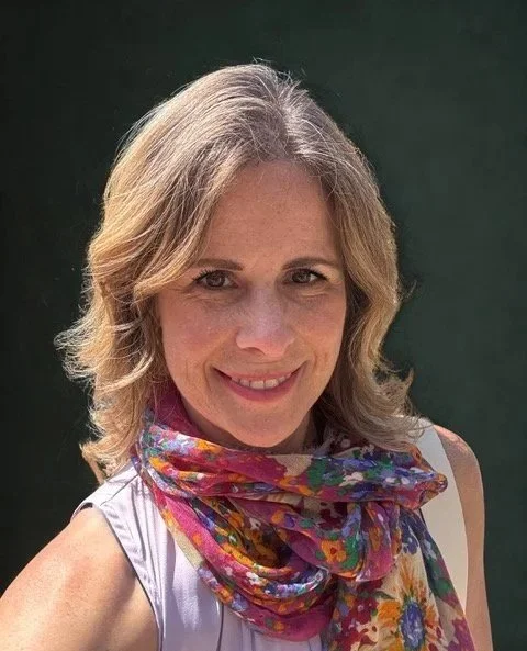 A smiling woman with shoulder-length blonde hair wearing a purple sleeveless top and a colorful floral scarf against a dark background.