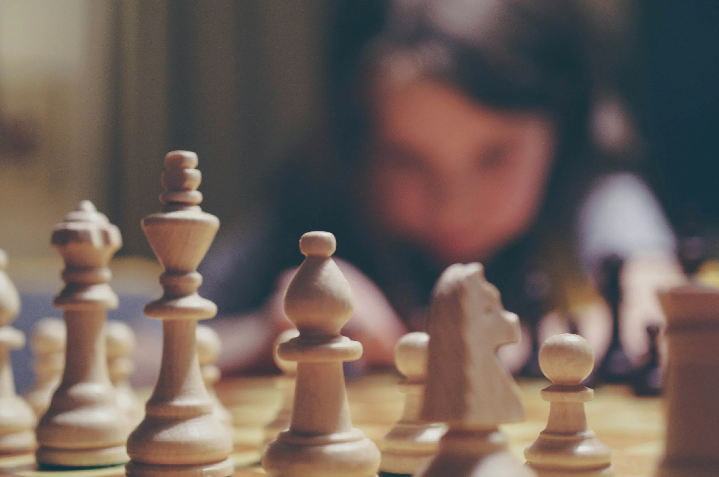 Close-up of a chessboard with wooden chess pieces in focus, and a blurred person in the background looking at the game.