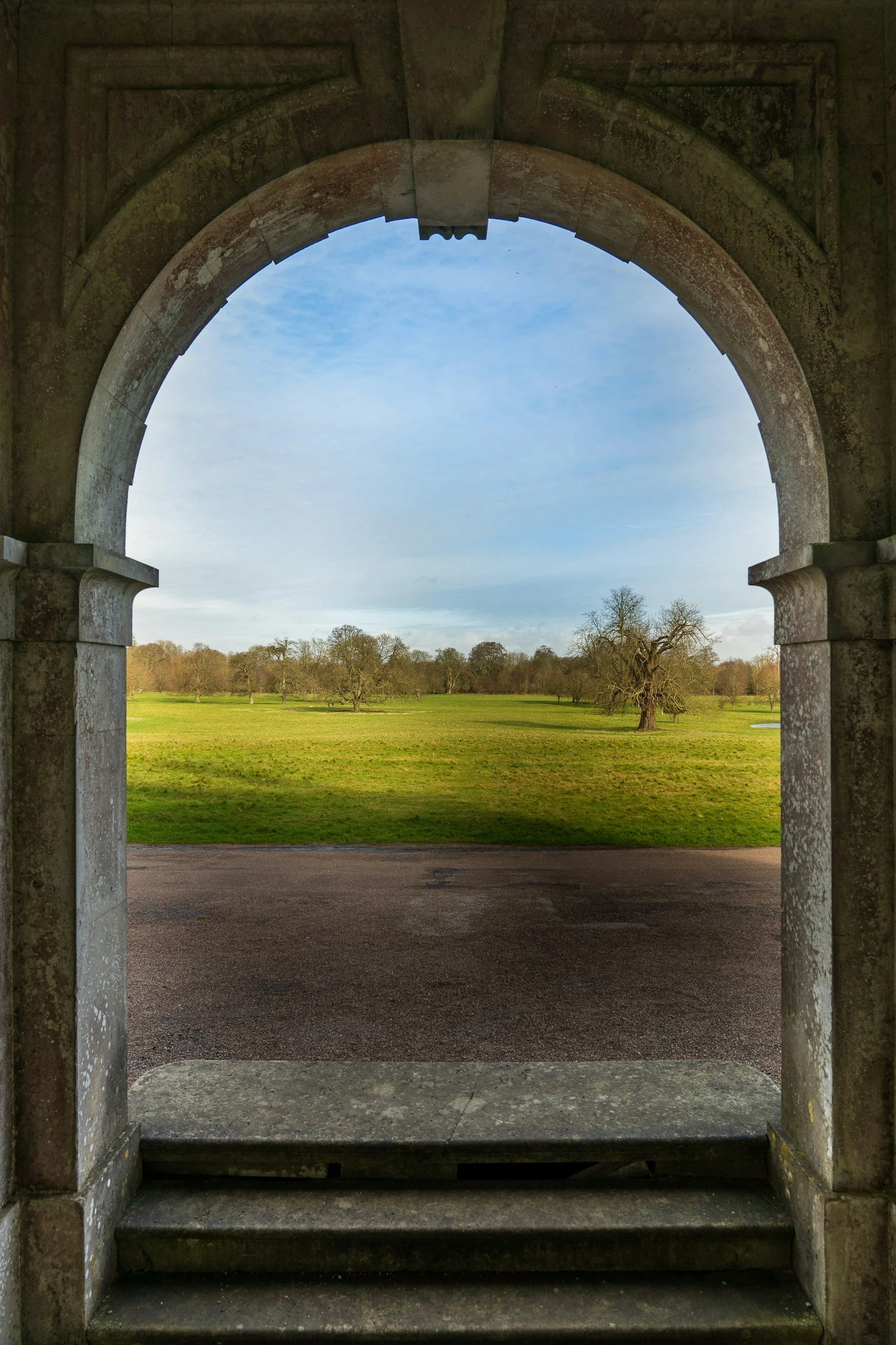 Stone archway framing a view of a grassy field with trees under a blue sky.
