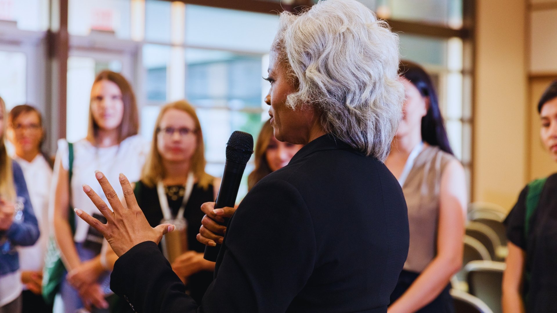 An elderly woman with gray hair holding a microphone and speaking to a group of women gathered in a bright indoor space. The women are listening attentively.