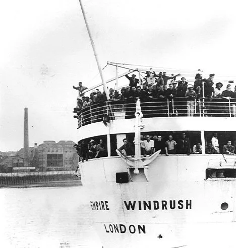 A black and white photo of a ship called the Empire Windrush arrives at harbor carrying a large group of men from different racial backgrounds.